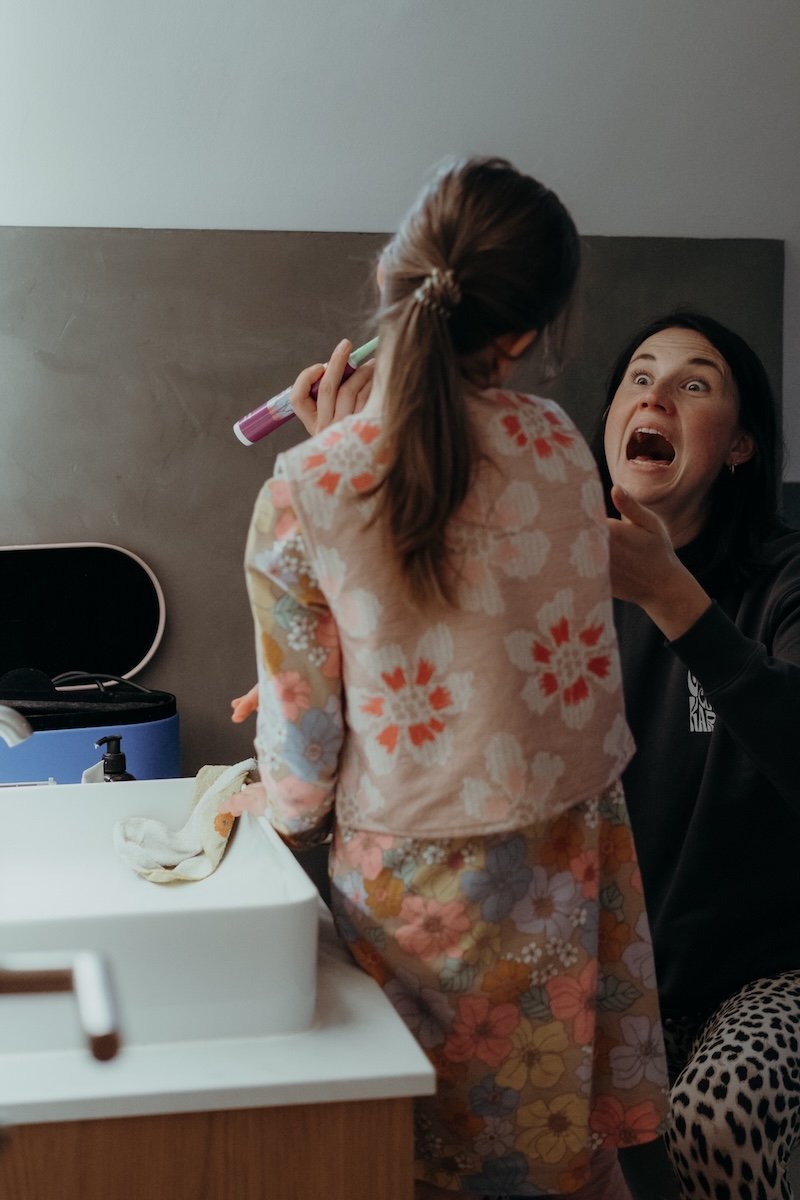 Eine Mama putzt ihrer Tochter, welche von hinten zu sehen ist, die Zähne. Die Mutter sieht die Tochter an und reißt den Mund weit auf.