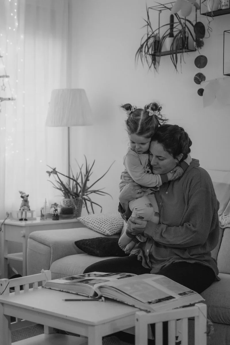 Eine Mama sitzt mit ihrer jungen Tochter im Arm auf dem Sofa, während beide zusammen ein Fotoalbum mit Kinderfotos anschauen.