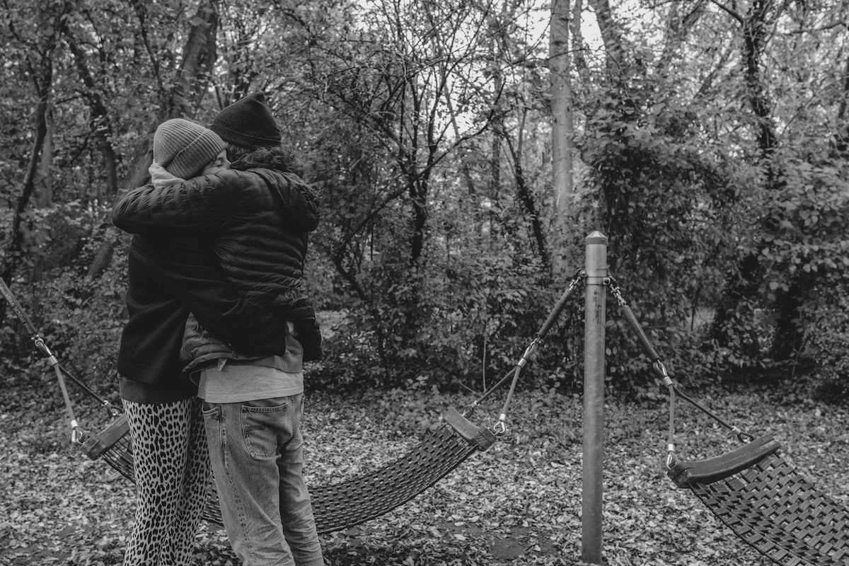 Die Eltern umarmen sich in einer ruhigen Minute der Zweisamkeit auf einem Spielplatz, im Hintergrund sind zwei Hängematten zu sehen.