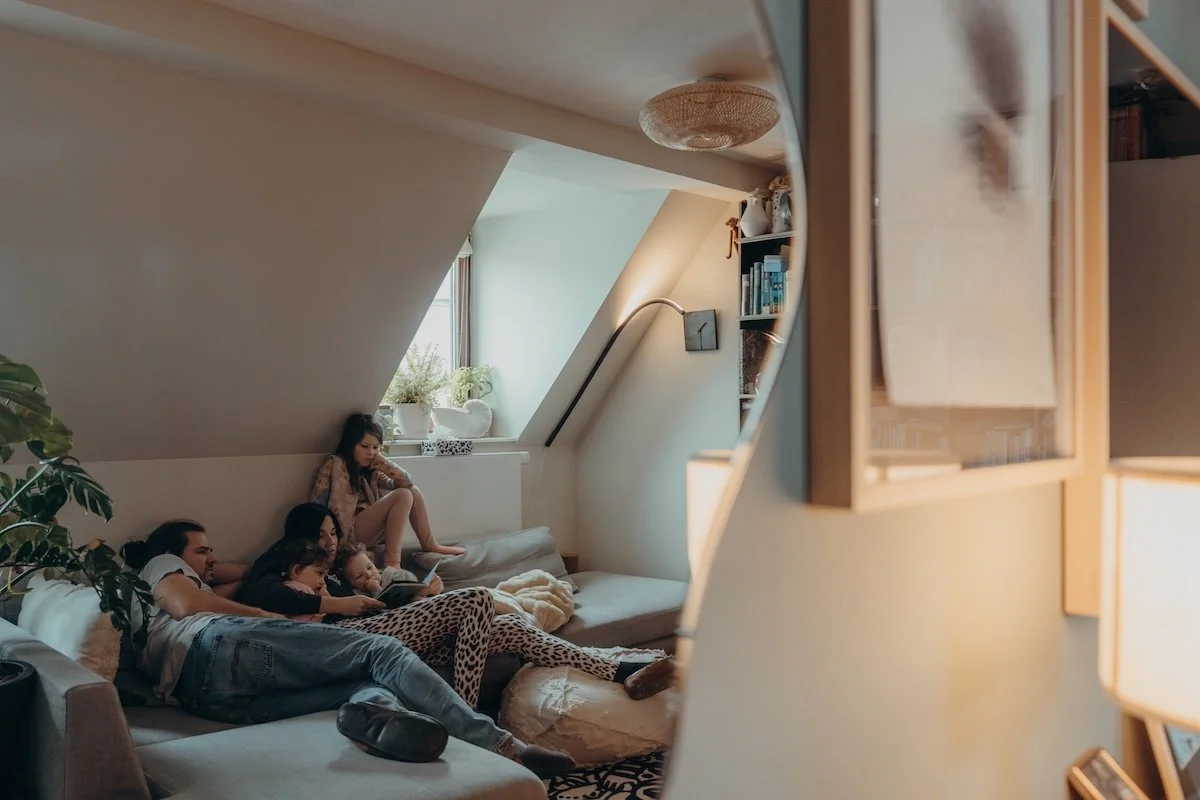 Eine Familie mit drei Kindern liegt entspannt auf den Sofa, während die Mama ein Buch vorliest. Die Szene spiegelt sich in einem Wandspiegel, daneben hängt ein Bilderrahmen aus Holz.