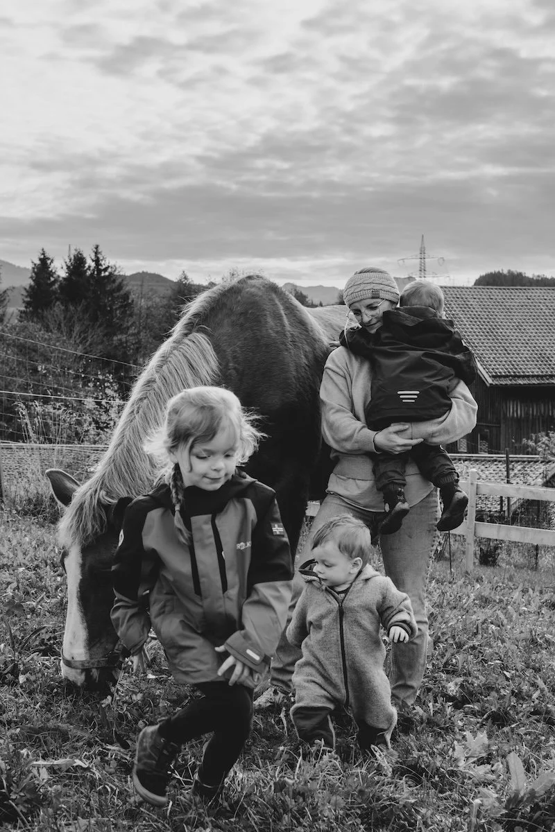 Eine Mama hält ihren Sohn im Arm, während ihre beiden anderen Kinder vor ihr laufen, neben ihnen steht ein Pferd.