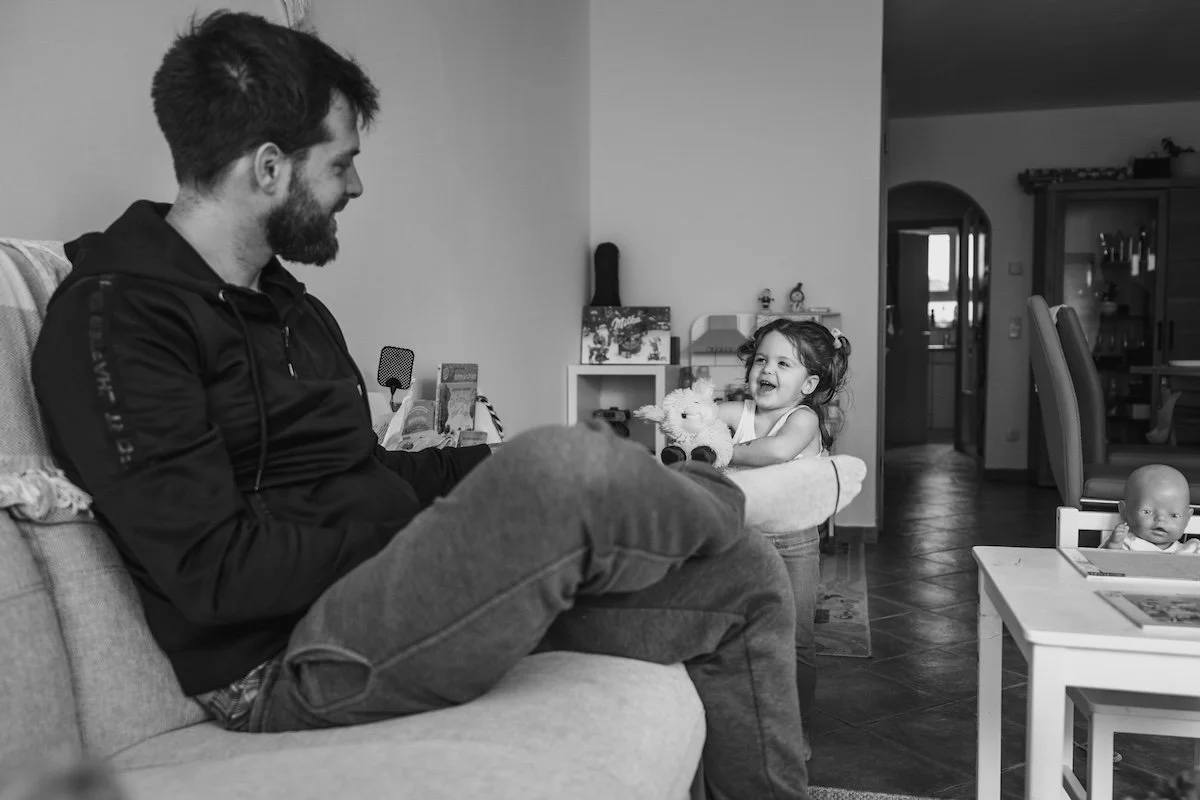 Der Papa sitzt auf dem Sofa, während er lächelnd zu seiner kleinen Tochter schaut, die ihm strahlend ein Kuscheltier entgegenhält.