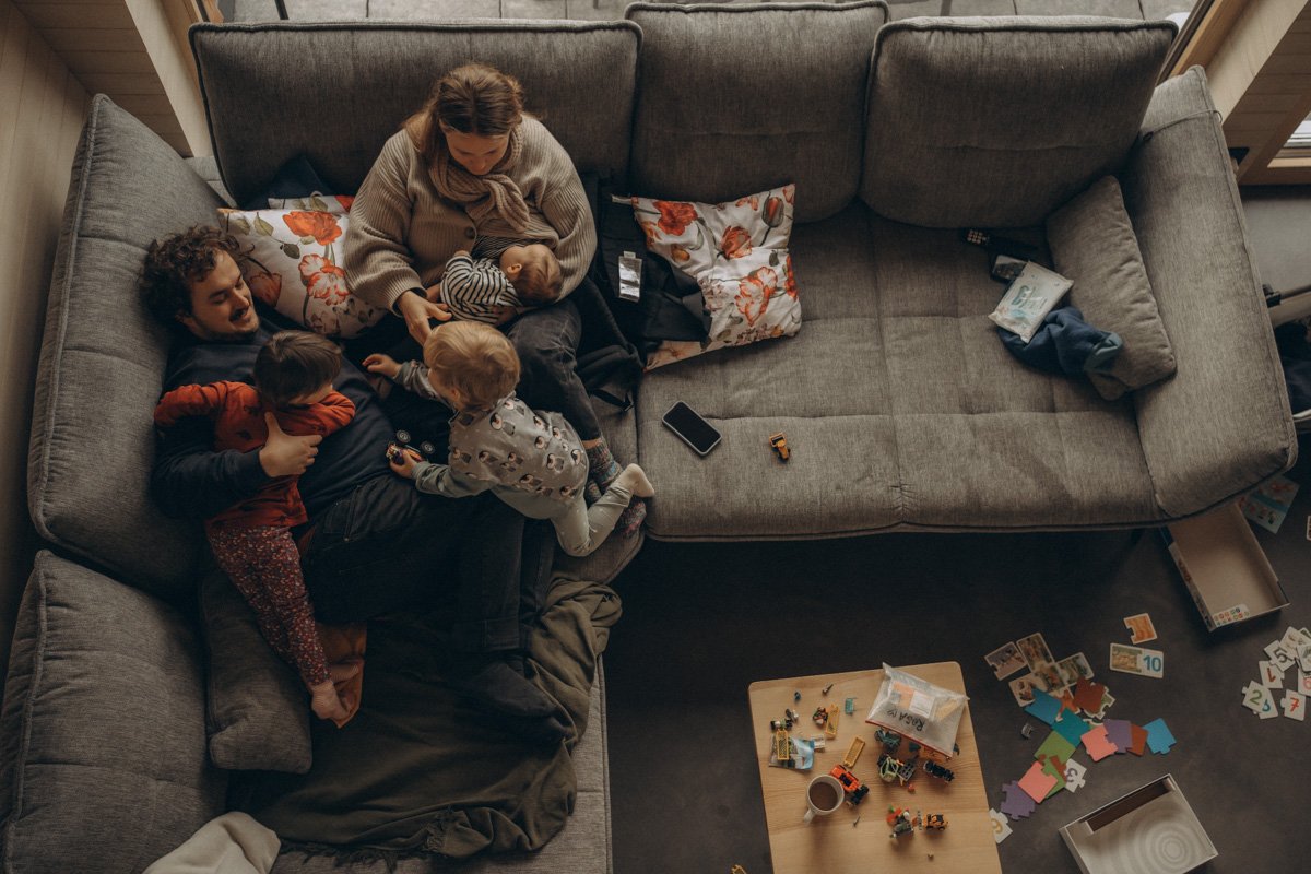 Fotoshootings im Wochenbett als Geschenkidee zur Geburt, über die sich jede Mama freuen wird. Eltern mit zwei Kleinkindern und einem Baby sitzen auf dem Sofa, überall Spielzeug auf dem Boden