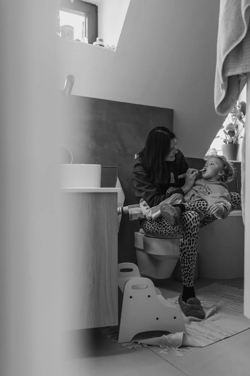 Eine Mama sitzt im Badezimmer und putzt ihrem kleinen Sohn die Zähne, während dieser auf ihrem Schoß sitzt.