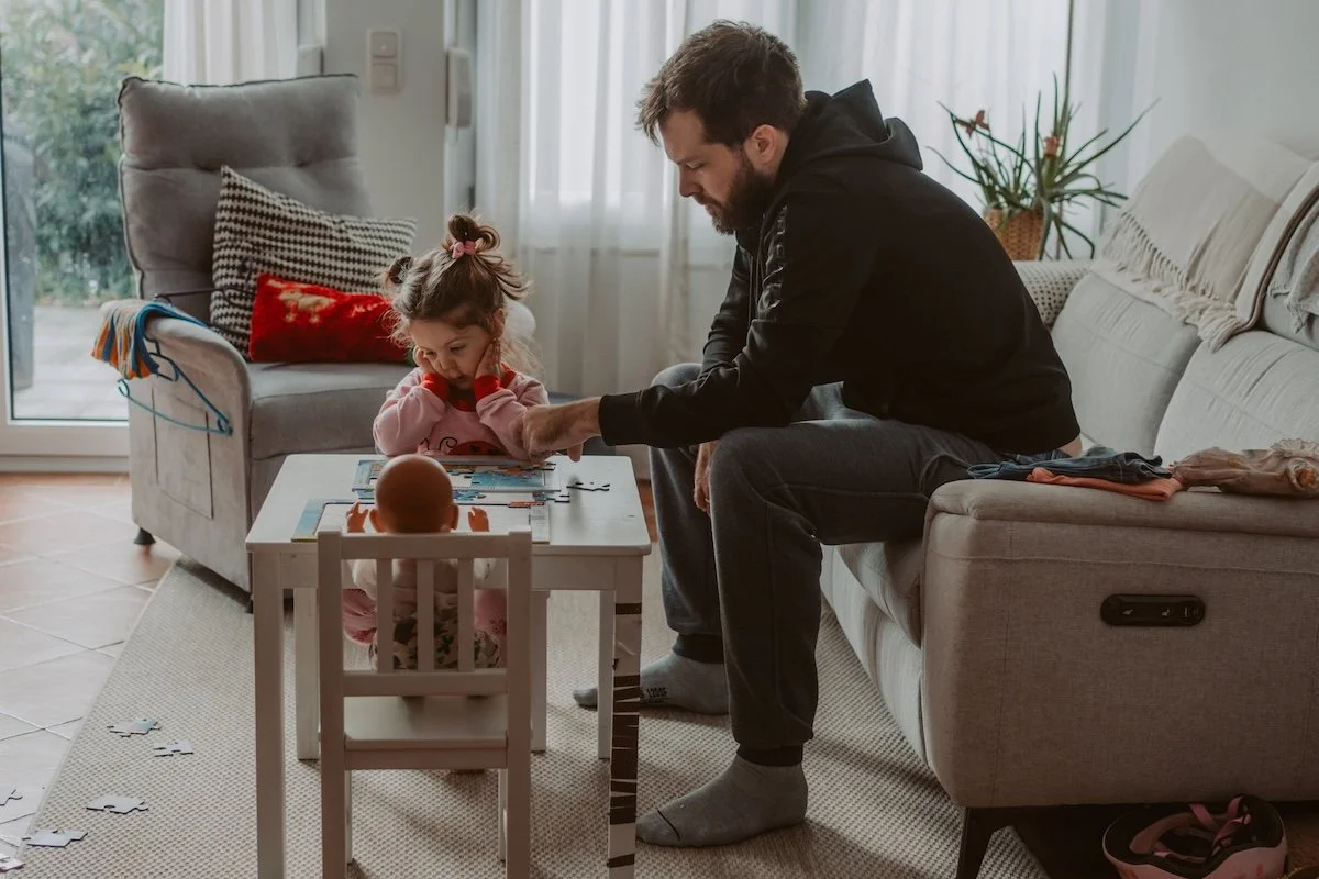 Tochter und Papa sitzen am Sofatisch und spielen ein Spiel, während eine Puppe auf einem Kinderstuhl ebenfalls am Tisch sitzt und dabei zusieht.