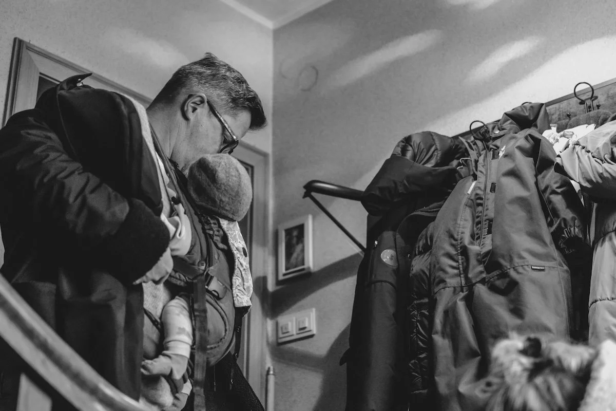Ein Papa zieht sich im Treppenhaus die Jacke an, während seine neugeborene Tochter in einer Trage an seinem Bauch sitzt.