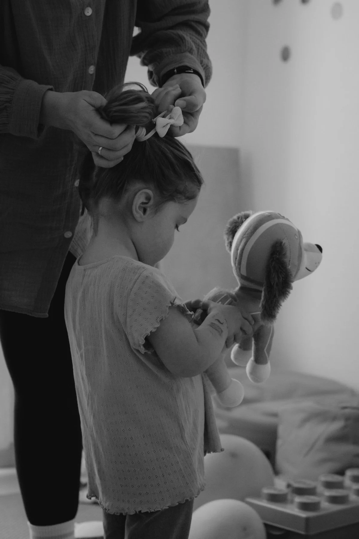 Ein junges Mädchen spielt mit ihrem Stoffhund in der Hand, während seine Mama ihr einen Zopf bindet.
