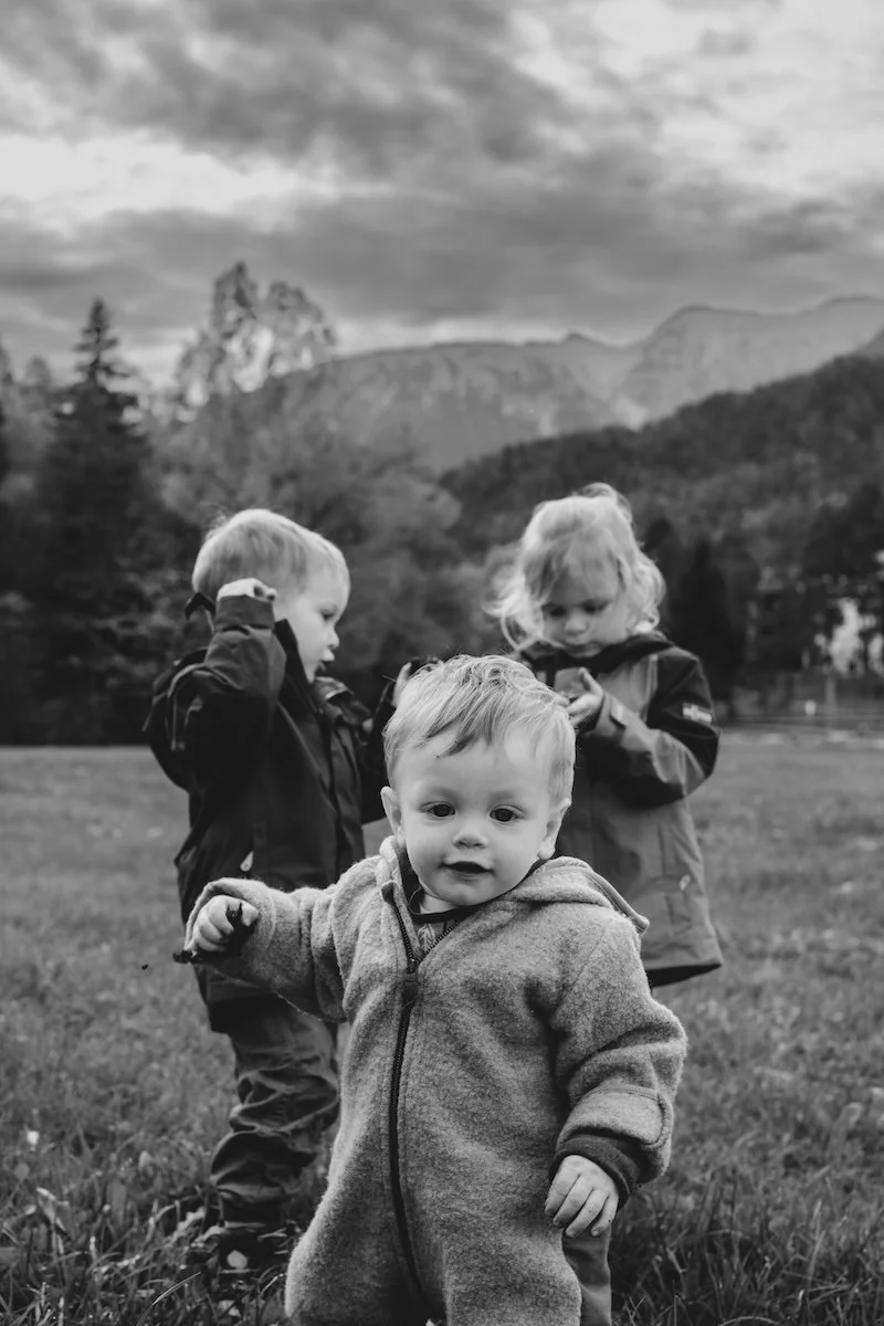 Drei Geschwister spielen auf einer Wiese in den Bergen, das jüngste Kind ist im Vordergrund, der ältere Bruder steckt sich eine Blume hinter das Ohr, die ältere Schwester begutachtet die Blume in ihrer Hand.