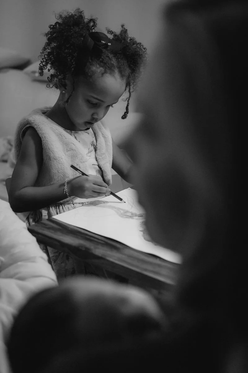 Ein junges Mädchen malt ein Plazentabild, während die Mama im Vordergrund die kleine Schwester im Arm hält.