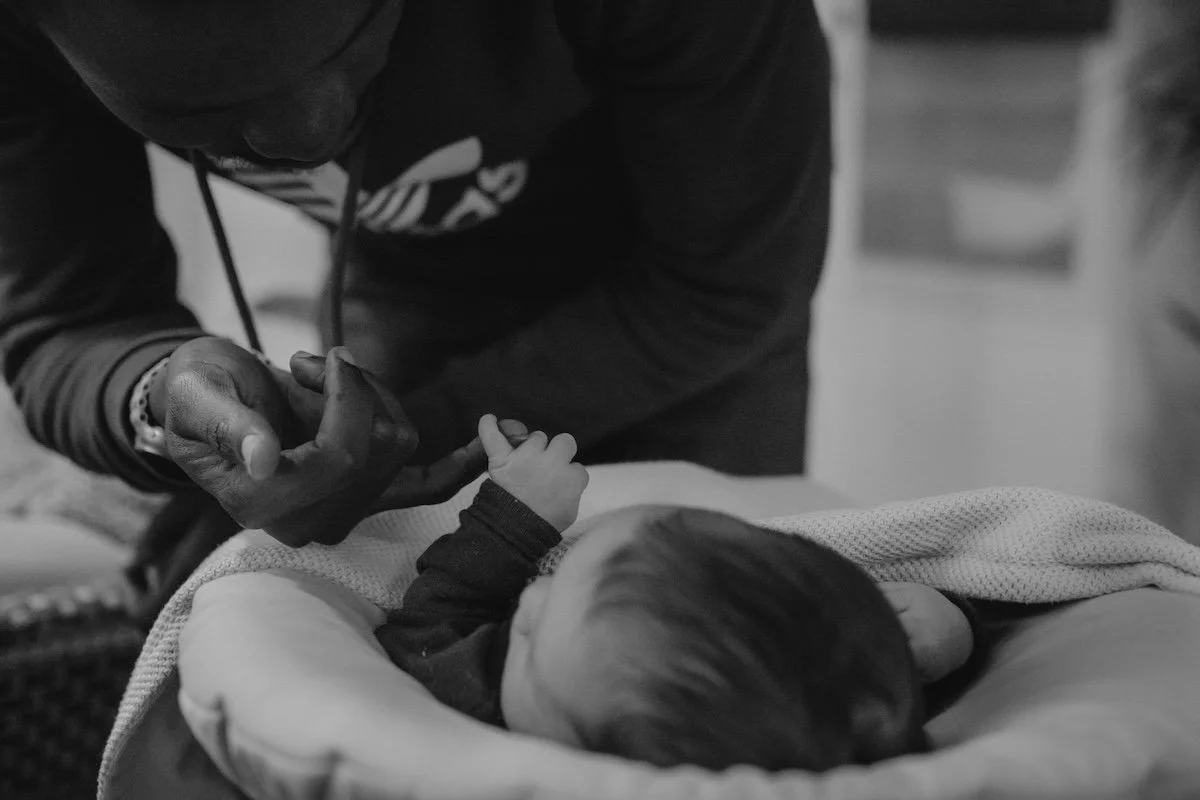 Der Papa beugt sich zu seiner neugeborenen Tochter, welche in einem Bettchen liegt und mit ihrer kleinen Hand den großen Finger des Papas umschlingt.