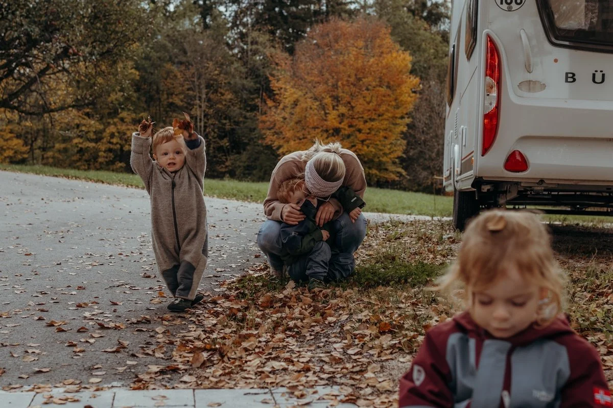 Eine Mama hält ihren Sohn tröstend im Arm, während ihr anderer Sohn neben ihr Blätter in die Luft wirft und ihre Tochter mit einem Blatt auf dem Kopf ein Stück entfernt versunken im Spiel vor ihr sitzt.