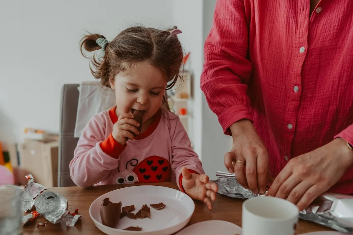 Ein junges Mädchen mit zwei Zöpfen isst Schokolade im Schlafanzug, während die Mama daneben steht und die Schokolade auspackt.