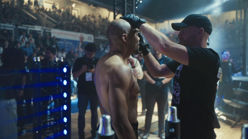 A fighter with a shaved head is getting prepared by a coach or medic in a large event venue with a crowd watching.
