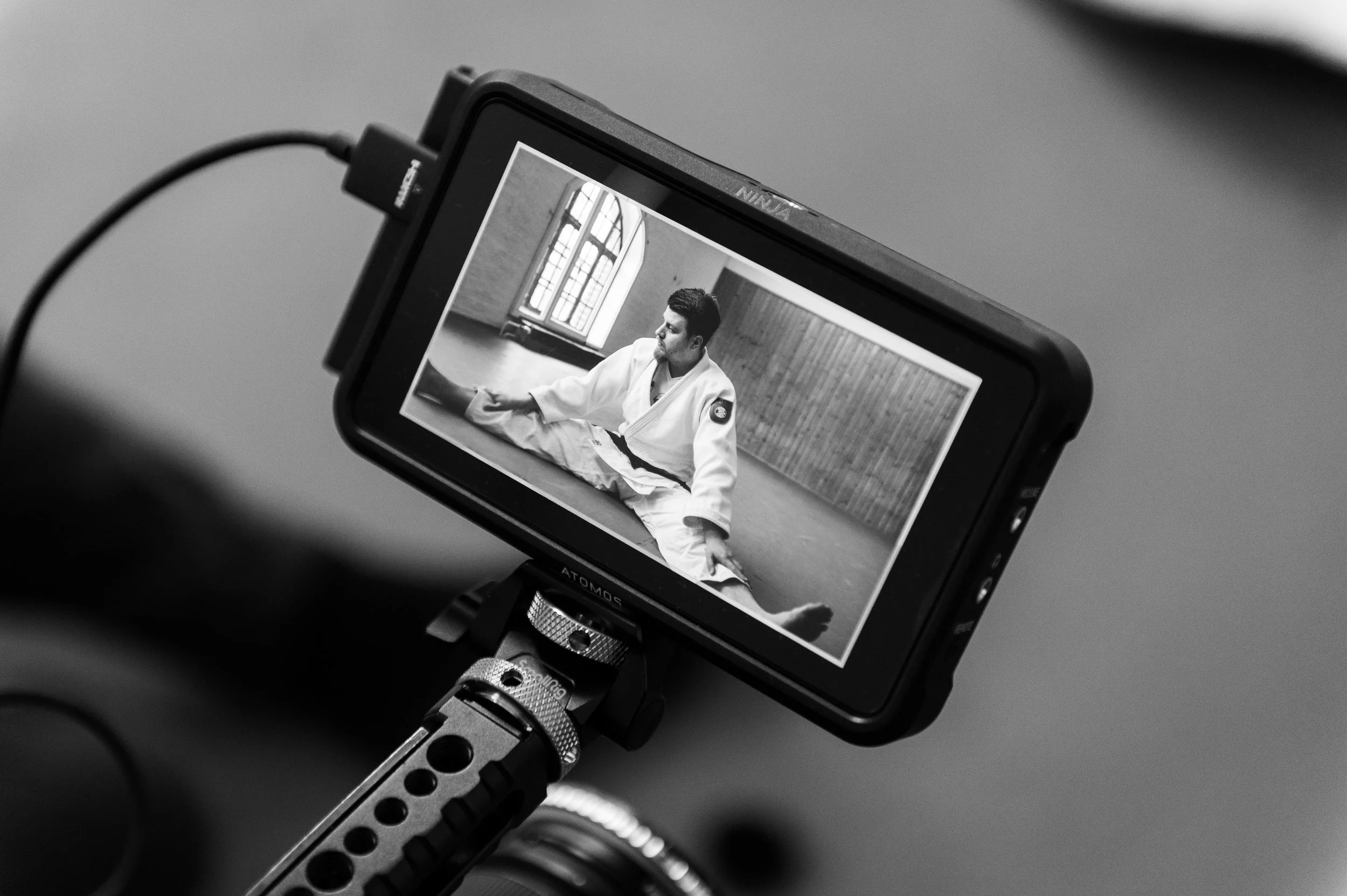 A camera screen displays a person in a white martial arts gi practicing a technique in a room with large arched windows and wooden paneling.