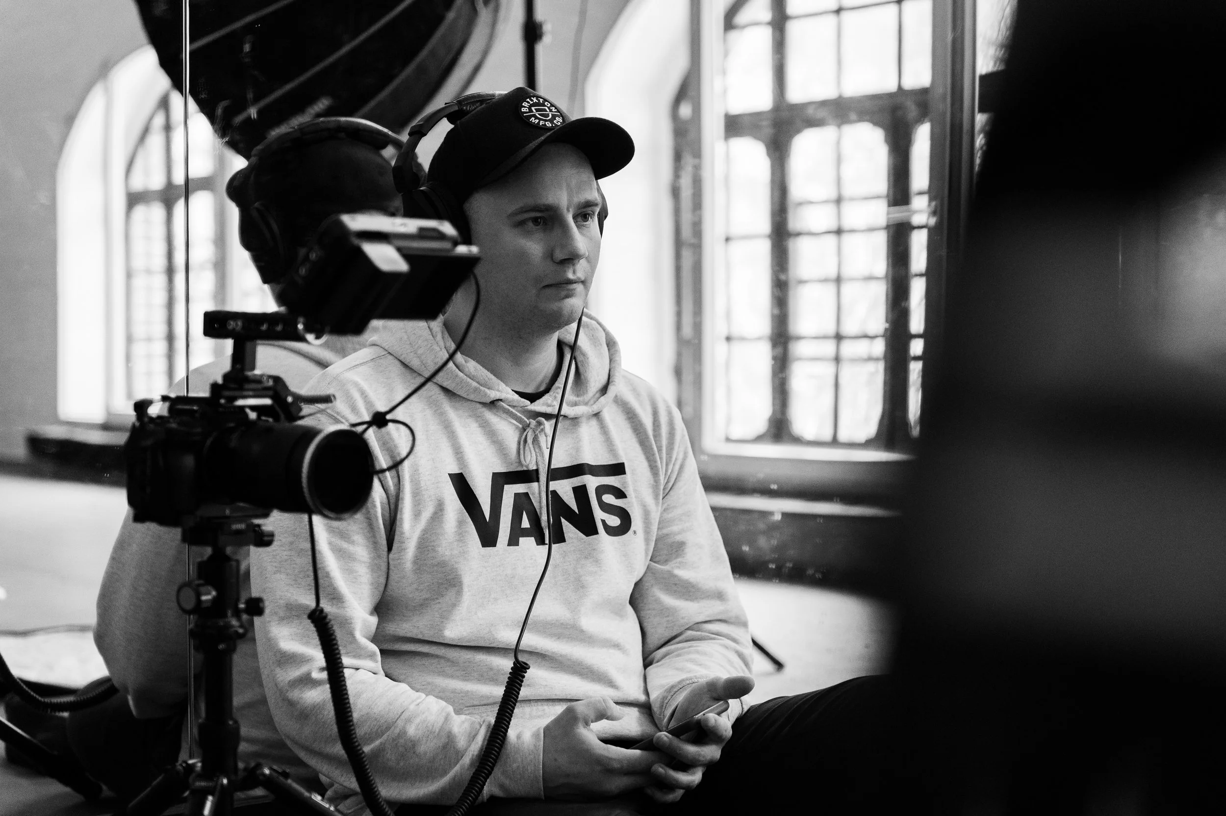 A young man with a cap, headphones, and a hoodie sitting indoors, looking at his phone, with a video camera setup nearby.