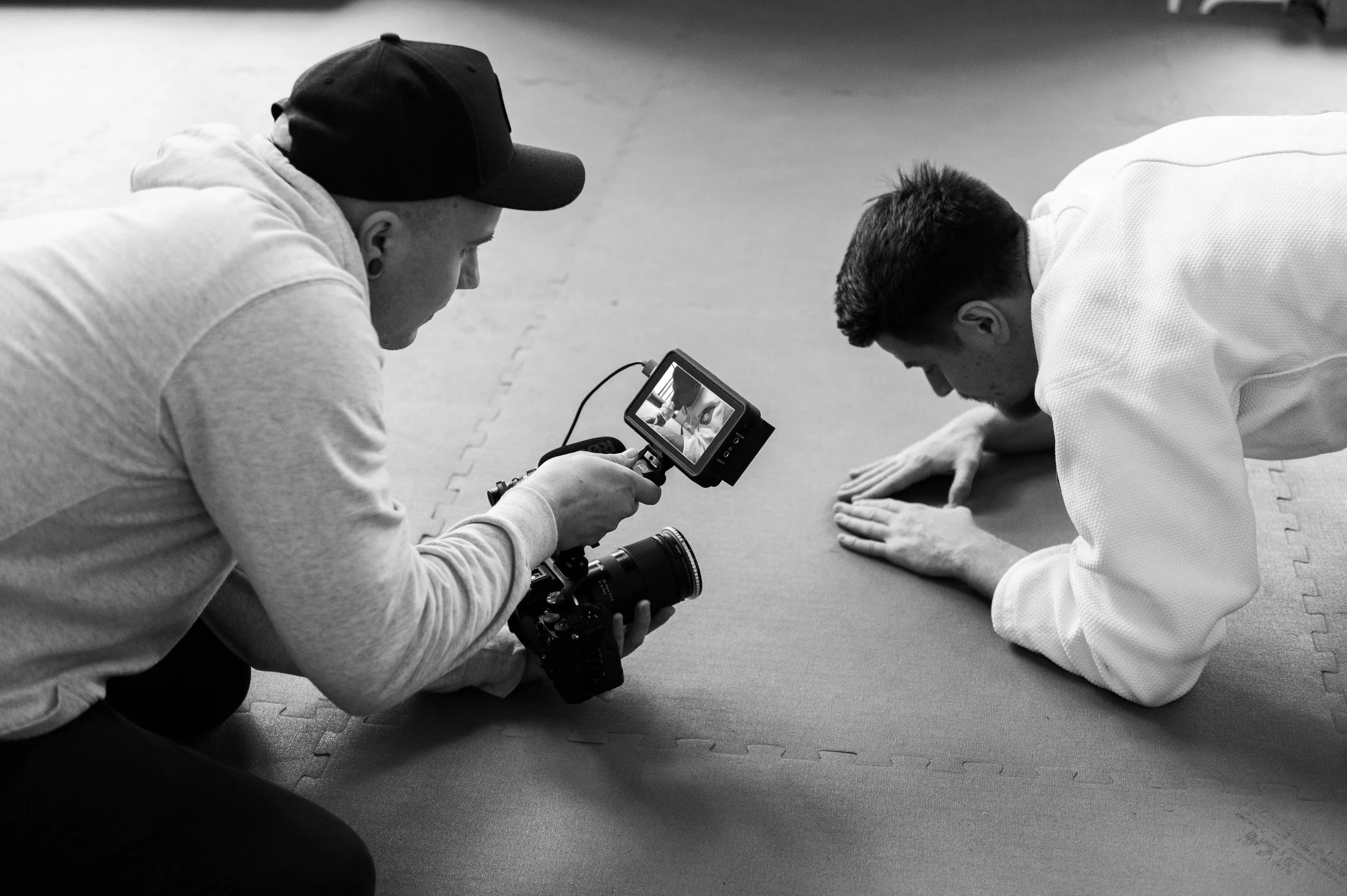 Two individuals with short dark hair, one with light skin and the other with medium skin, practicing a martial arts move on a padded floor. The person on the right, wearing a white martial arts gi, is positioned on their hands and knees, while the other person on the left, wearing a hoodie and a cap, is kneeling and holding a camera, filming or photographing the scene.