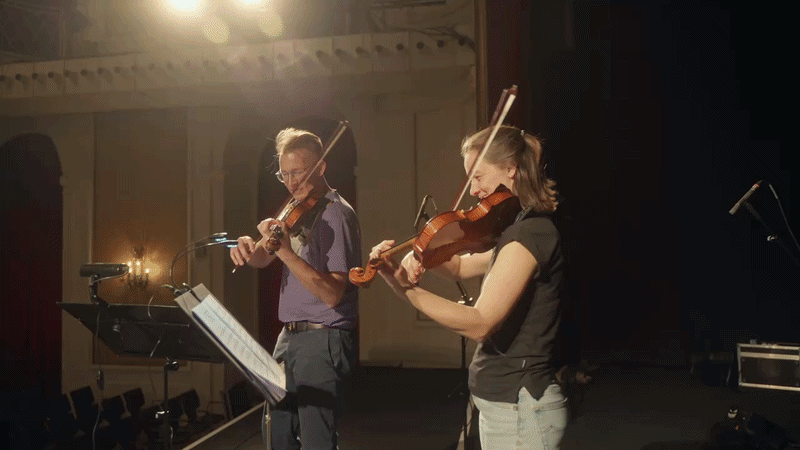 Two musicians playing violins on stage during a performance, with sheet music and stage lighting.