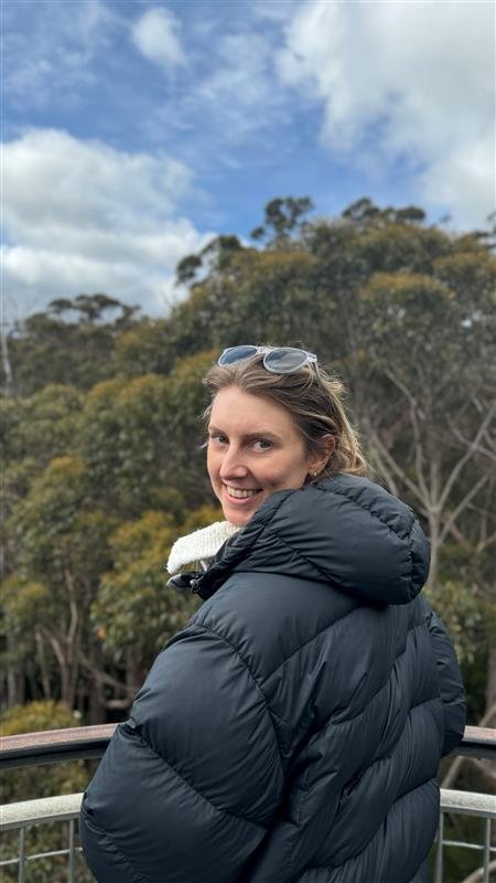 A smiling woman in a black puffer jacket standing outdoors on a balcony with trees and a blue sky with clouds in the background, helped by a Career Coach.