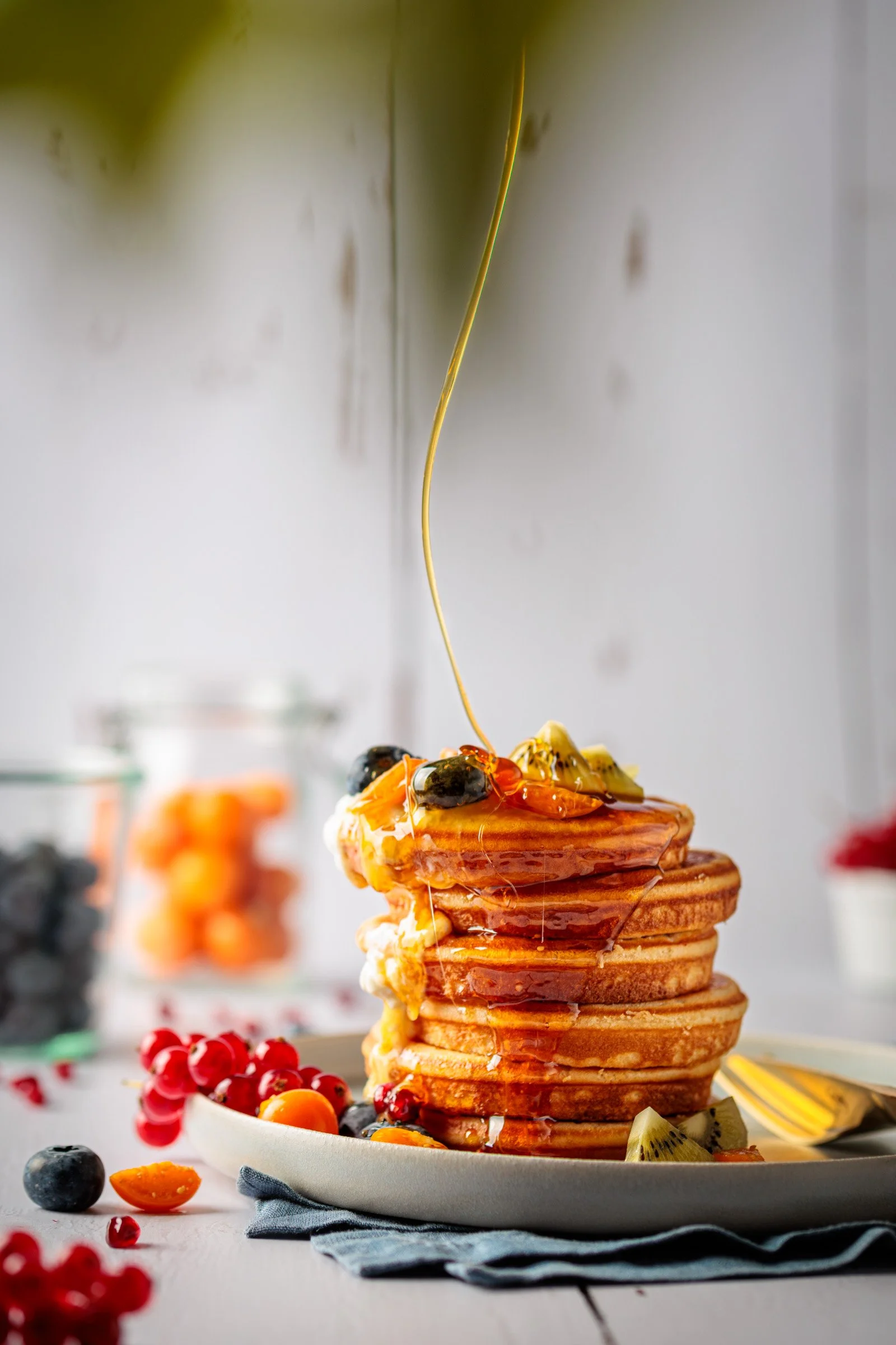 Haufen von goldbraunen Pfannkuchen mit Ahornsirup, frischem Obst wie Kiwi, Heidelbeeren, Johannisbeeren und Passionsfrucht, auf einem weißen Teller