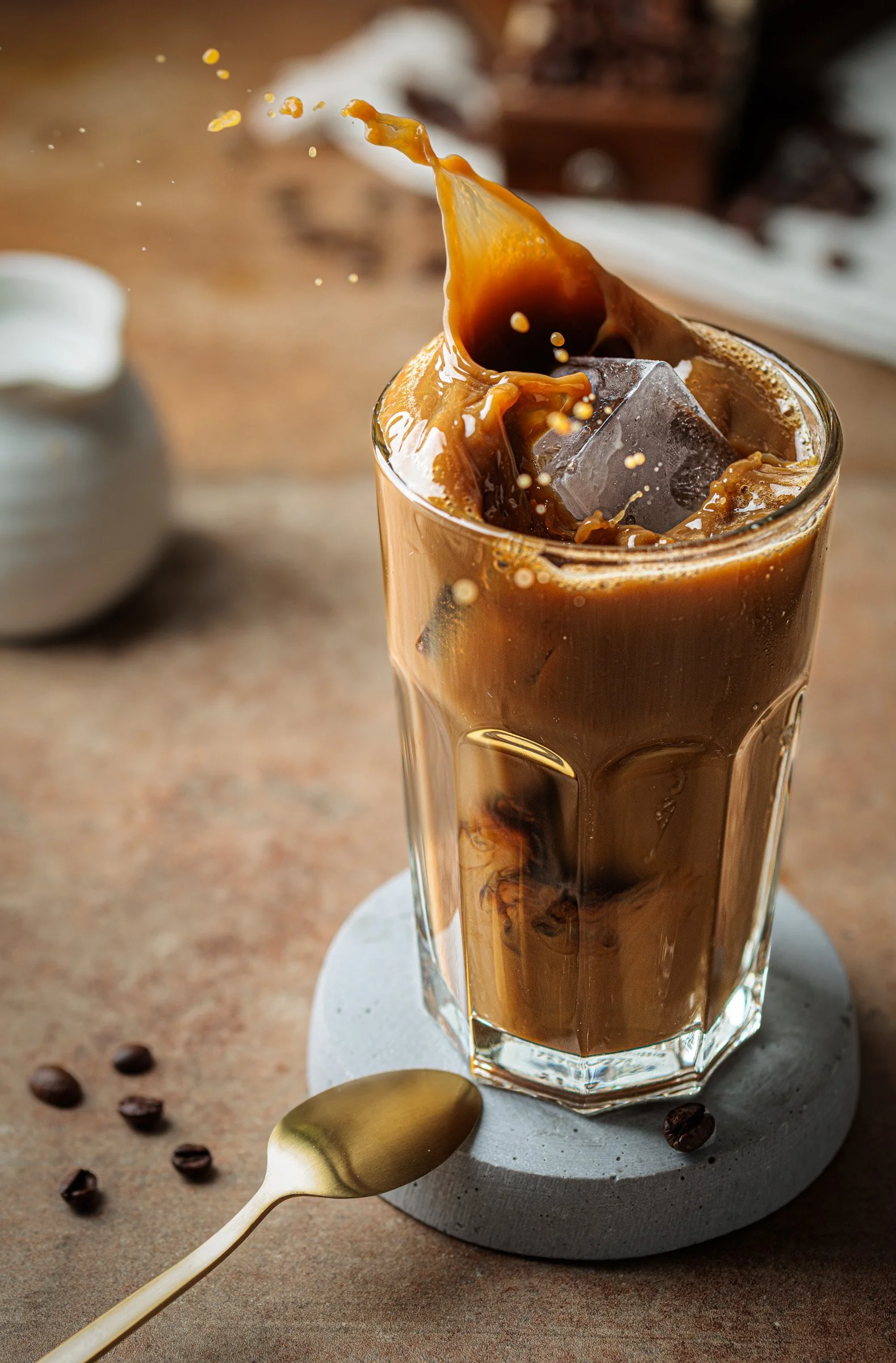 Ein Glas Eiskaffee mit Eiswürfeln und spritzenden Kaffee. Ein goldener Löffel liegt neben dem Glas auf einem runden Steinuntersetzer, umgeben von Kaffeebohnen, auf einem Tisch.