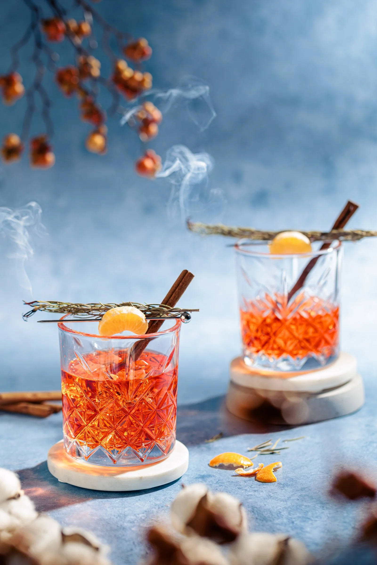 Zwei Gläser mit süßen Aperitifs, garniert mit Zitronenscheiben und Zimtstangen, auf blauer Tischplatte mit weiteren Zutaten und Dekorationen.