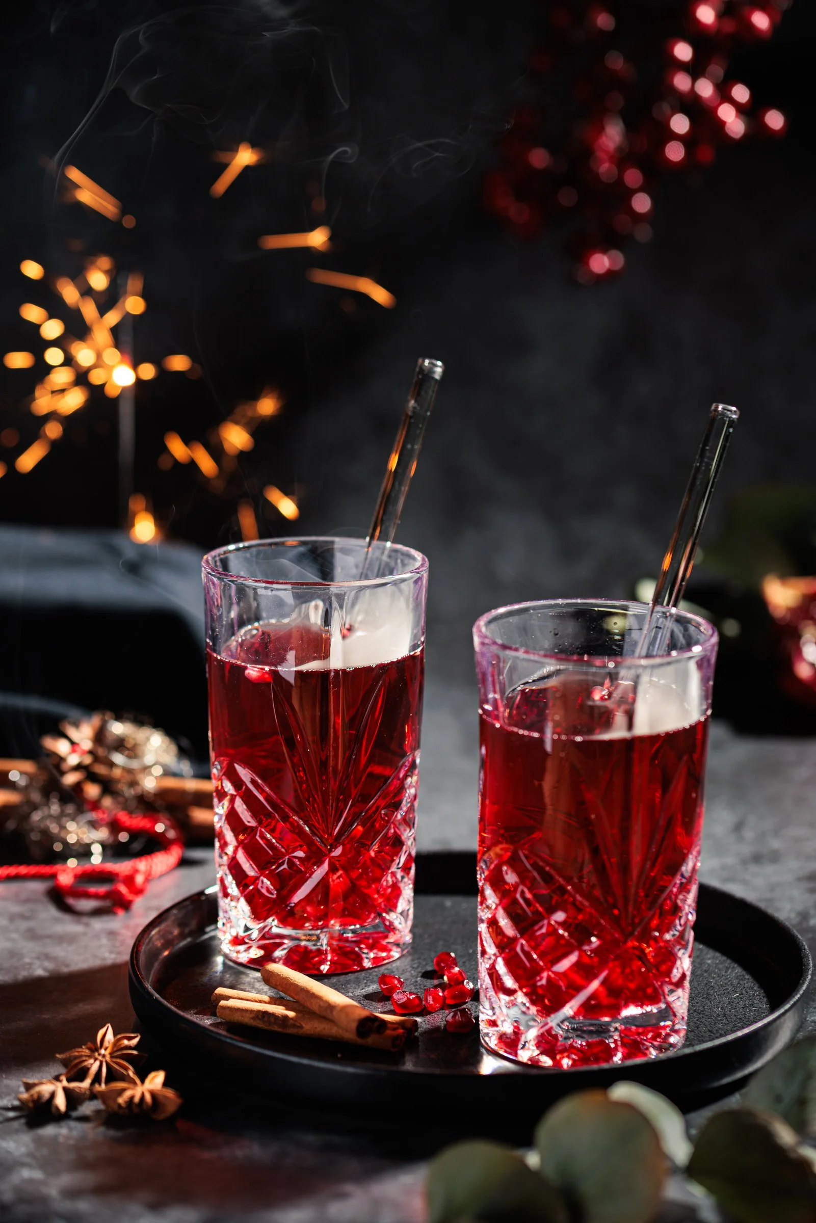 Zwei Kristallgläser mit roten Getränken, Strohhalme, Zimtstangen und Granatapfelkerne auf einem schwarzen Tablett, festliche Dekoration und Feuerwerk im Hintergrund.