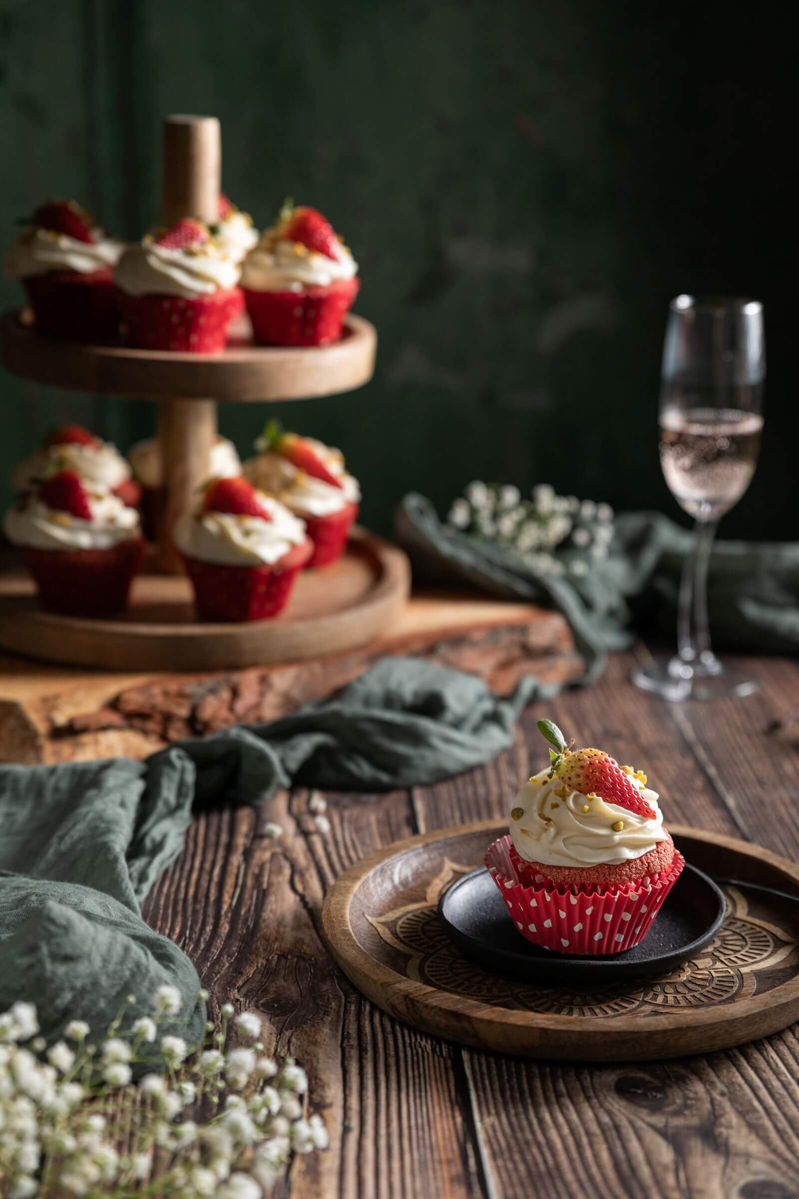 Ein einzelner Cupcake mit weißer Creme, Erdbeere und goldenen Zuckerdekorationen auf einem Holztablett im Vordergrund, weitere Cupcakes auf einem dreistufigen Holzständer im Hintergrund, ein Glas Sekt und weiße Blumen auf dem Tisch.