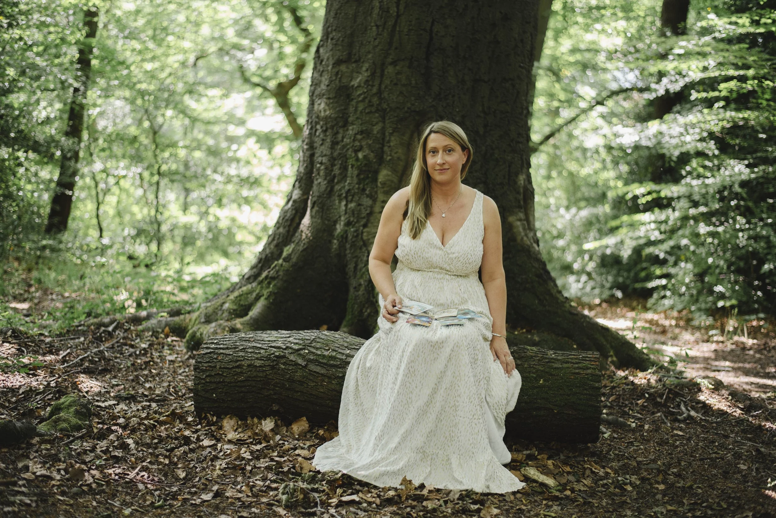 A woman in a white dress sitting on a fallen tree trunk in a lush green forest.
