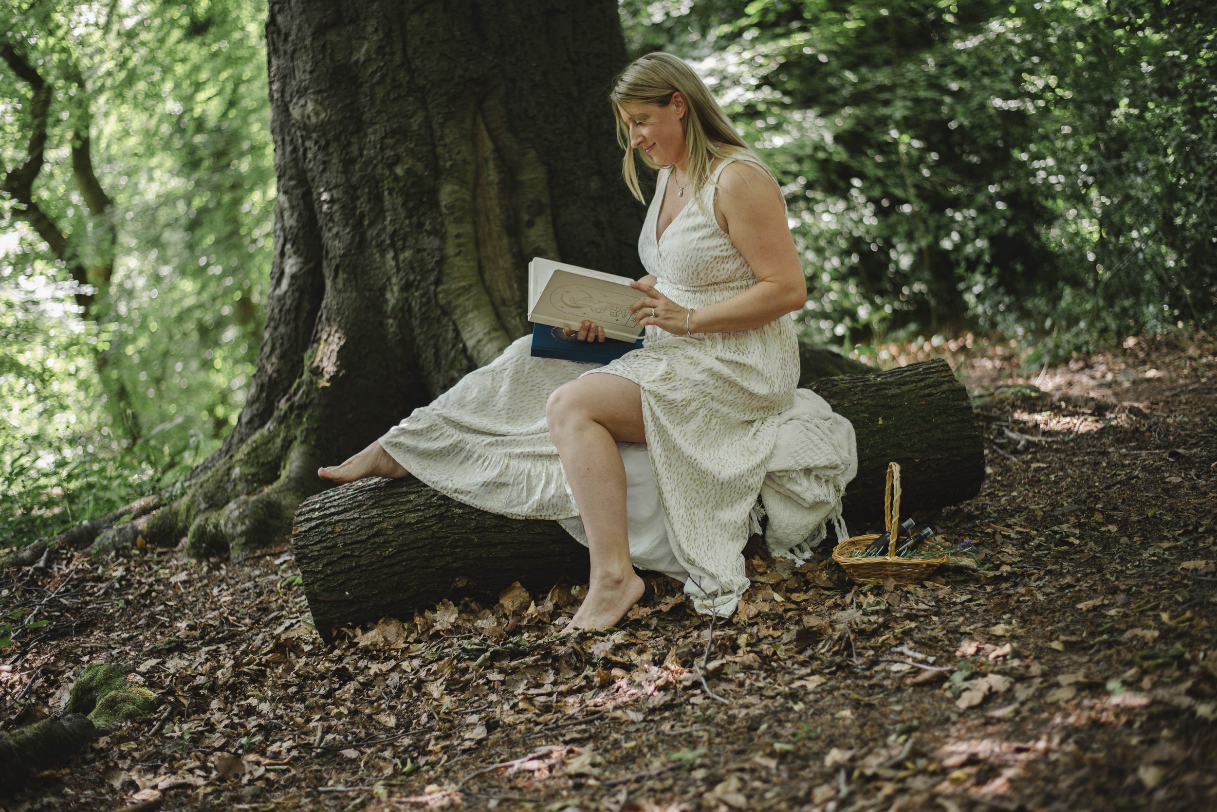 A woman in a white dress sitting on a tree log in a forest, looking at a book, with a basket on the ground beside her.