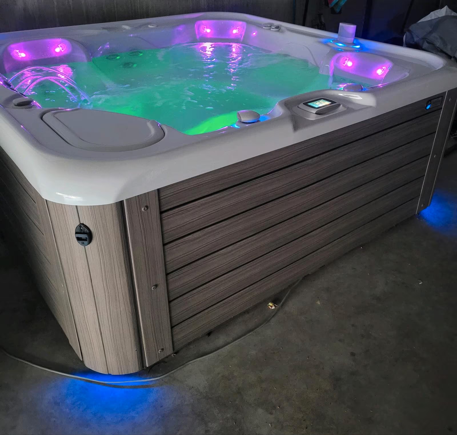 A hot tub with water illuminated by purple and green lights, set on a concrete floor with a wood-paneled exterior, and a digital control panel on the side.