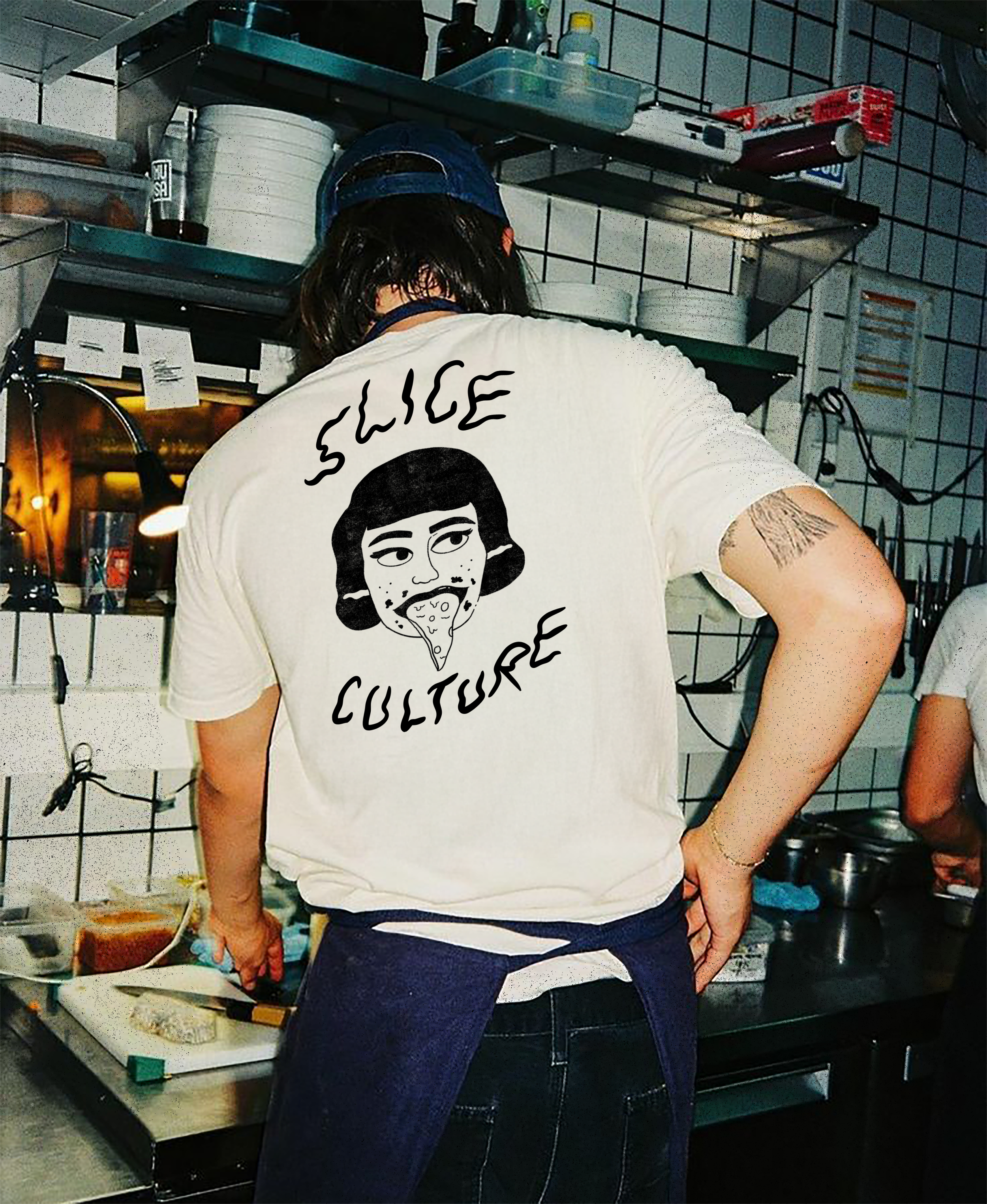 Person wearing a white t-shirt with a graphic of a woman eating pizza, and the words "Sliced Culture" printed on the back, standing in a kitchen preparing food.