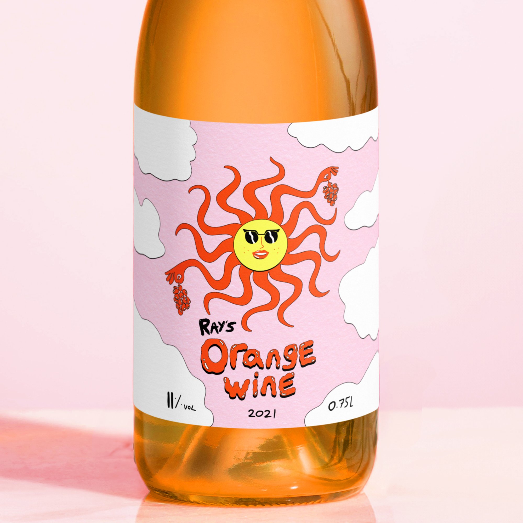 A bottle of orange wine with a pink label featuring a cartoon sun wearing sunglasses, surrounded by clouds and grape clusters, with the text 'Ray's Orange Wine,' '2021,' and volume 0.75 liters.