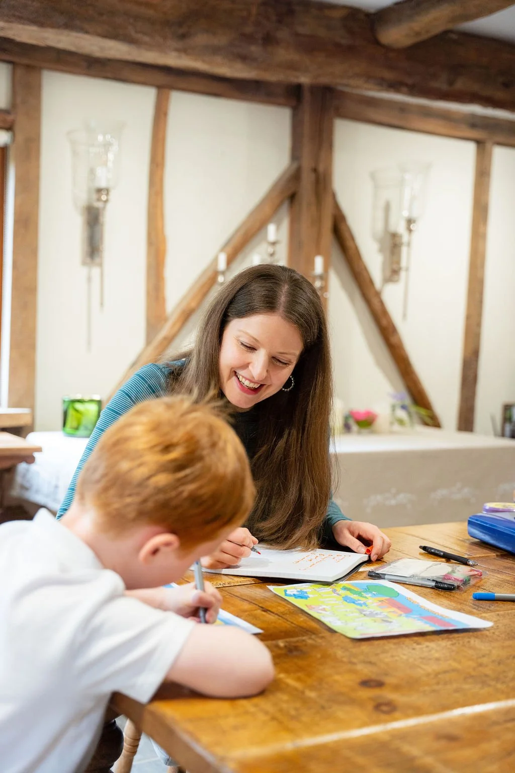 A woman and a young boy sitting at a wooden table engaged in an activity, with the woman smiling and the boy focused on drawing or writing. The room has rustic wooden beams and wall sconces.
