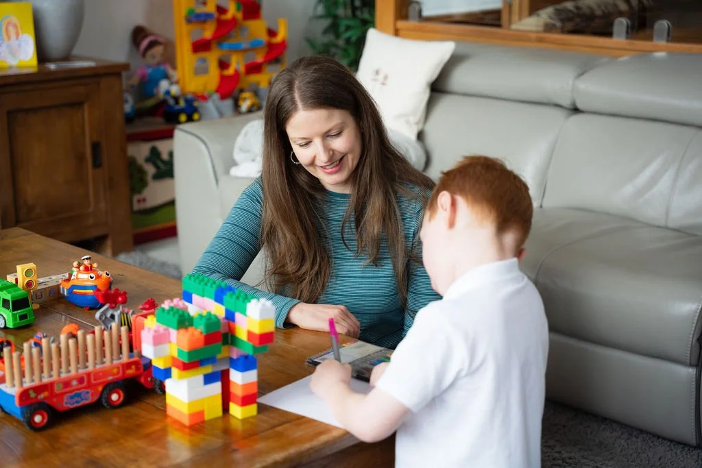 A woman and a young boy playing with colorful children's toys and building blocks at a wooden table in a cozy living room.