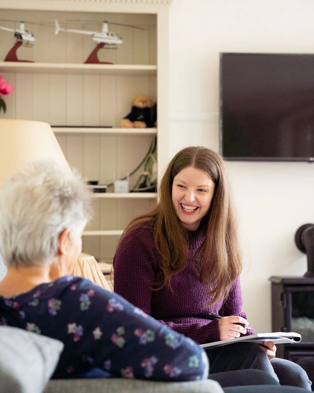 A woman with long brown hair smiling during a conversation with a woman with white hair, in a cozy living room.