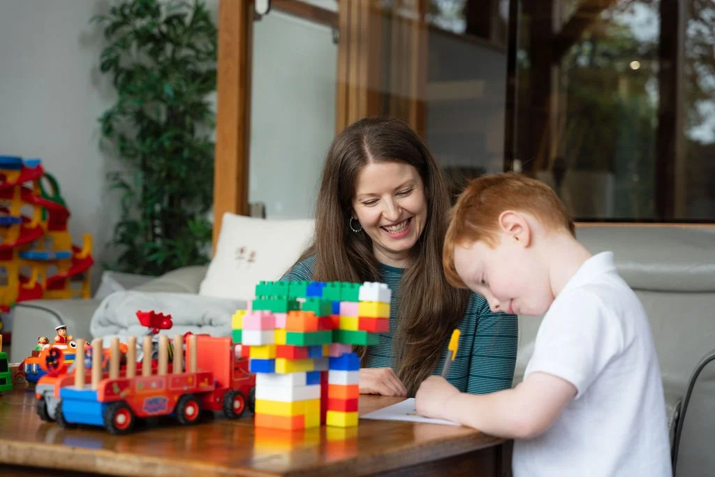 A woman and a young boy playing with colorful LEGO bricks on a wooden table. The woman is smiling while watching the boy as he builds. There are LEGO trains and structures on the table, and indoor plants and windows in the background.