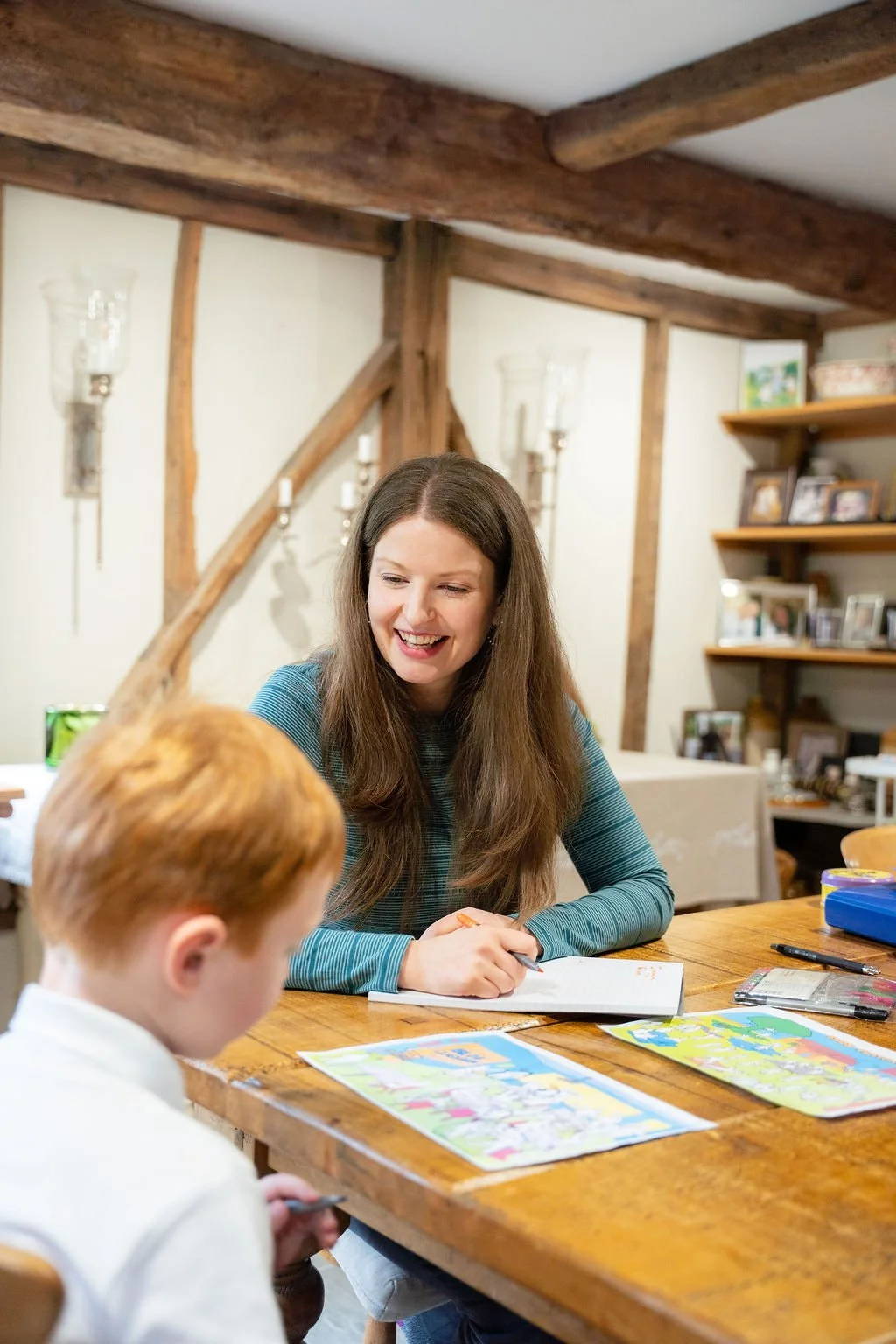 A woman with long brown hair smiling and engaging with a young boy with red hair at a wooden table. The table has colorful papers, a pen, and a pencil case. The room has rustic wooden beams and shelves with framed photos and decorative items.