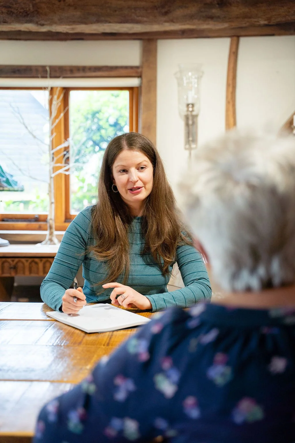 A woman with long brown hair wearing a green striped top sitting at a wooden table, engaged in conversation with a woman with short grey hair. The scene appears to be inside a cozy room with a window showing greenery outside.