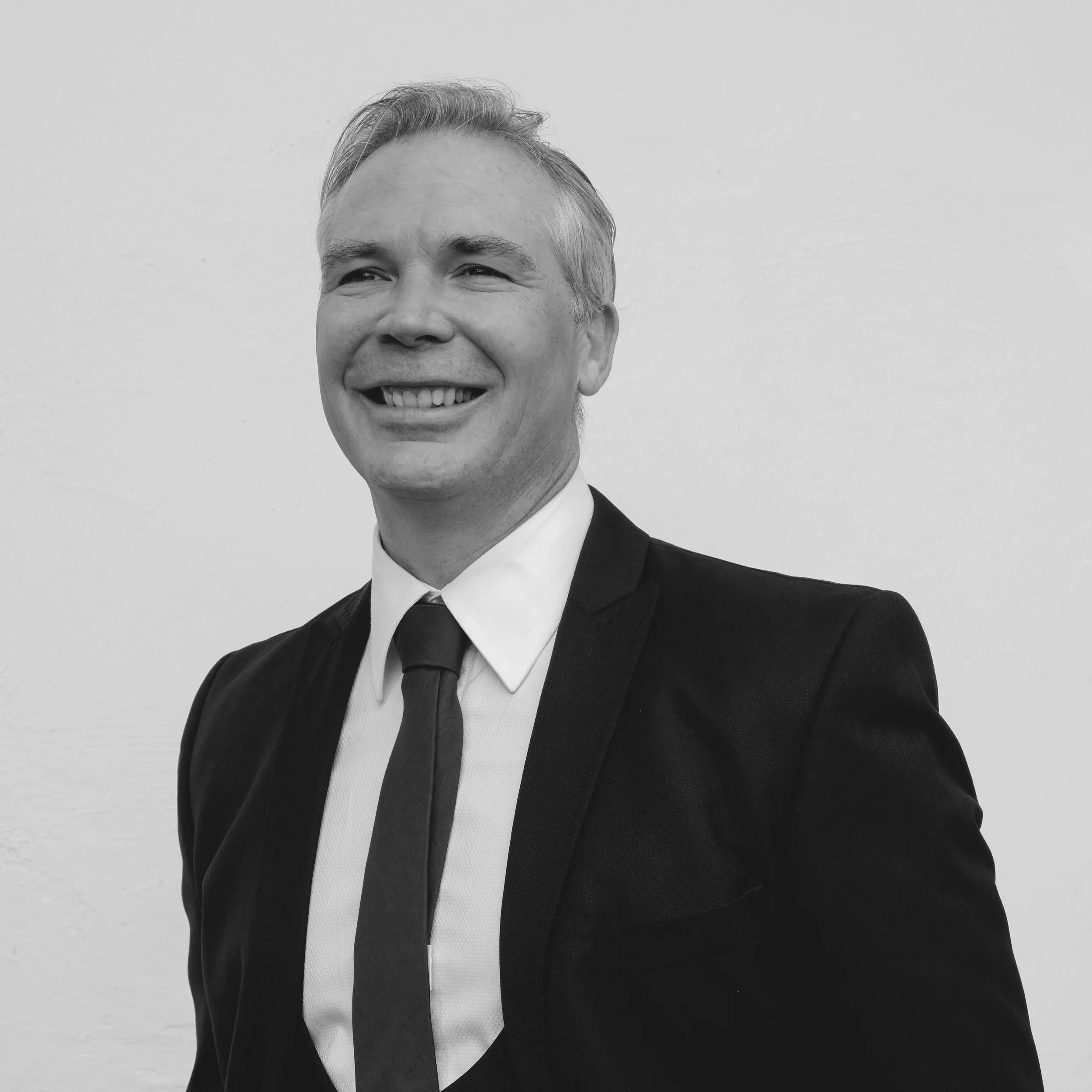 A man with short gray hair, wearing a white shirt and a watch on his left wrist, poses with his arms crossed against a black background.