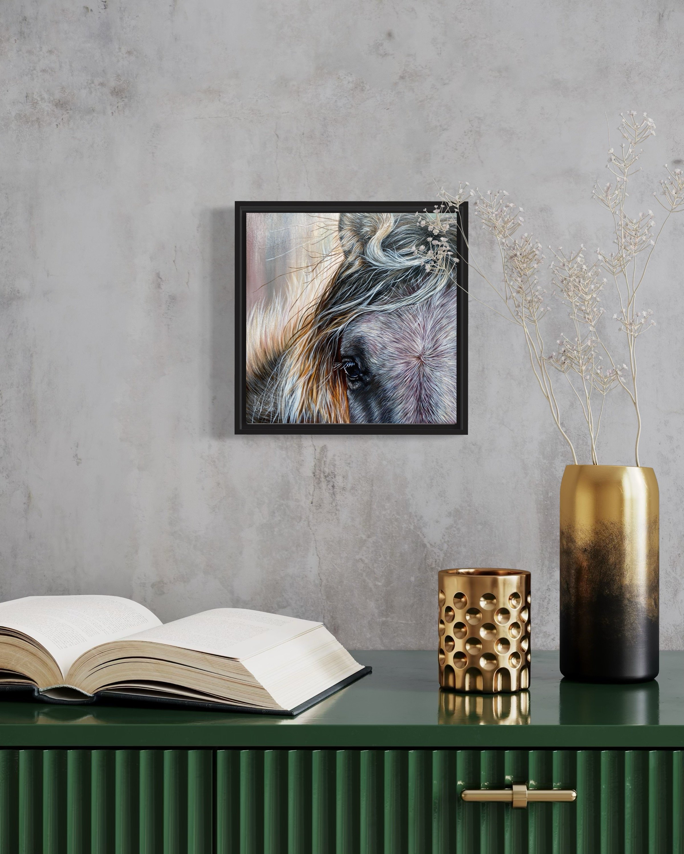 Ein Wandbild eines Pferdekopfes, ein offenes Buch, eine goldene Kerze mit Lochmuster und eine goldene Vase mit Trockenpflanzen auf einem grünen Sideboard vor einer Betonwand.