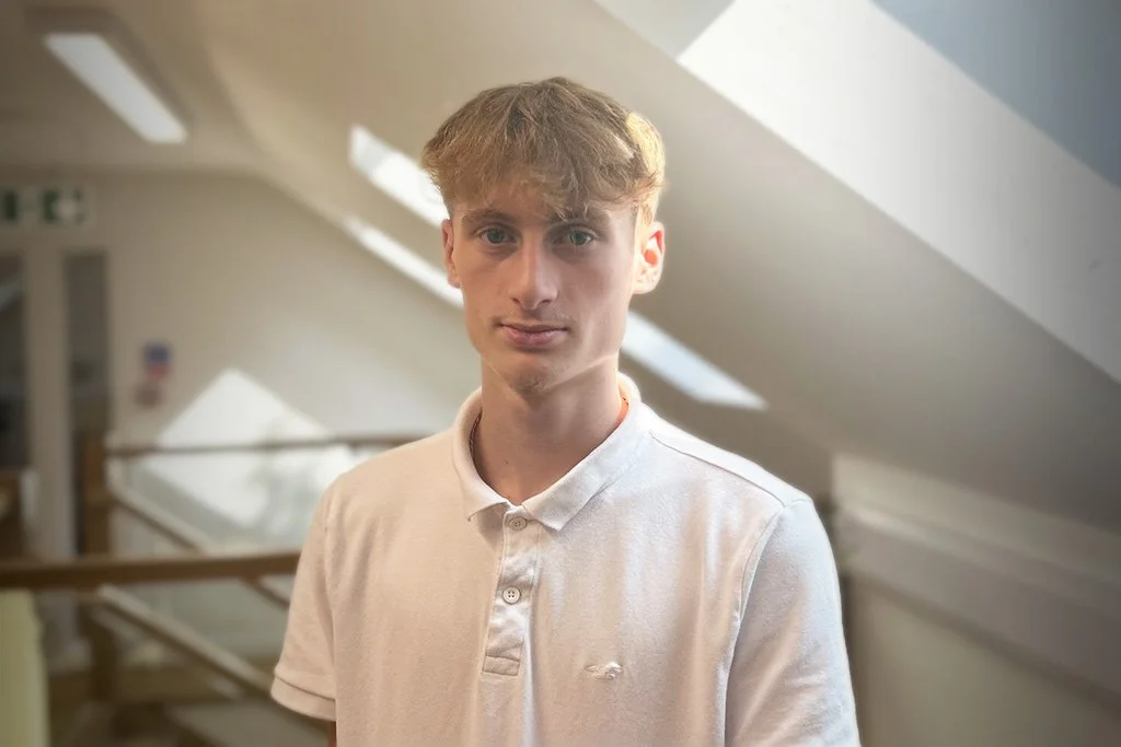 Young man in a white polo shirt standing indoors with natural light from windows. Background shows interior architecture with slanted ceiling and blurred details.