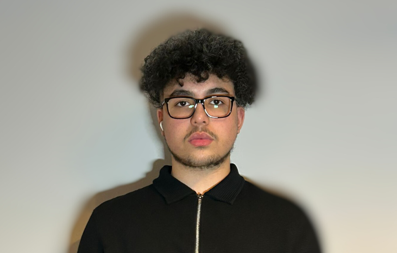 Person with curly hair and glasses wearing a black zip-up shirt against a plain background.