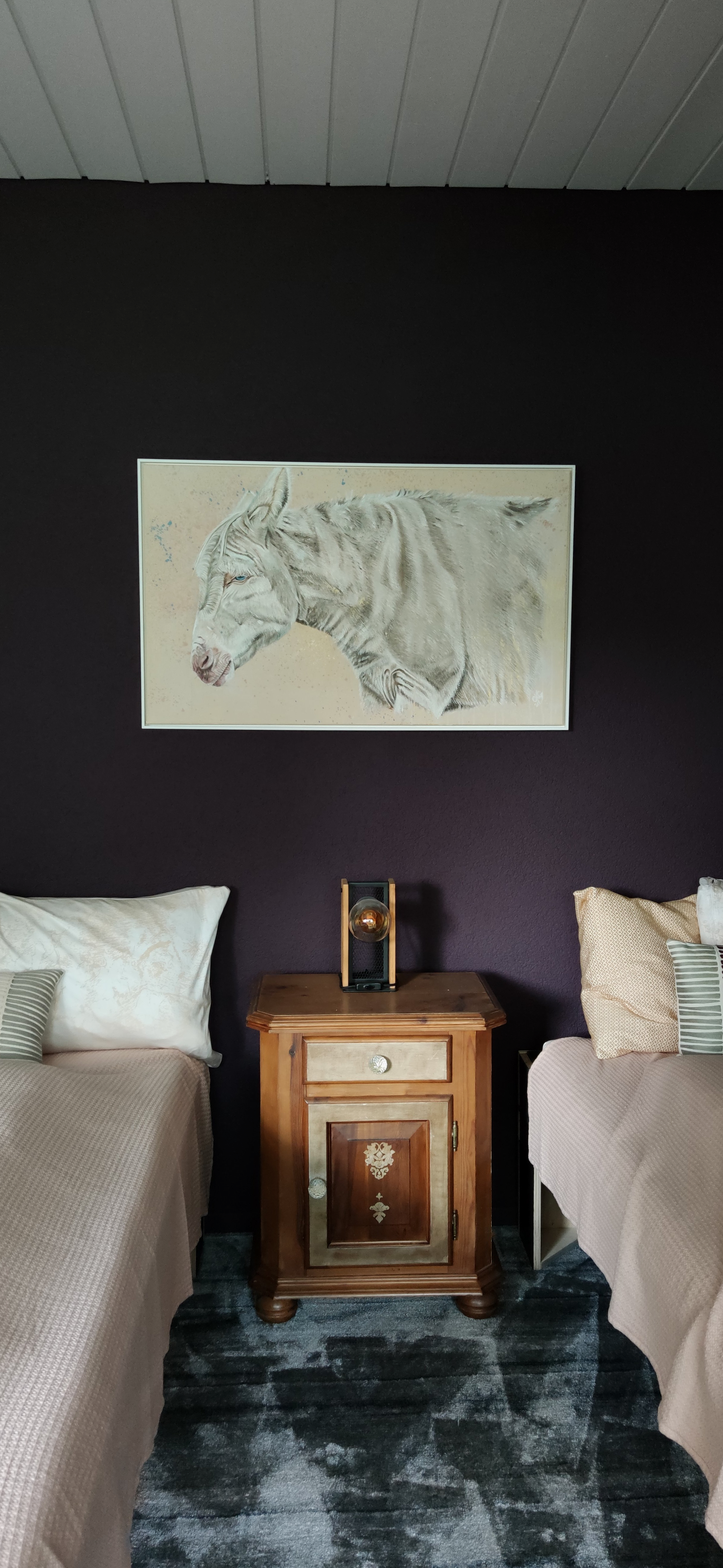 Ein Wohnzimmer mit einer dunkelvioletten Wand, an der ein Gemälde eines weißen Esel hängt. Zwischen zwei Betten steht ein Holz-Nachtisch mit einer Glühbirne in einer goldfarbenen Fassung auf der Oberfläche.
