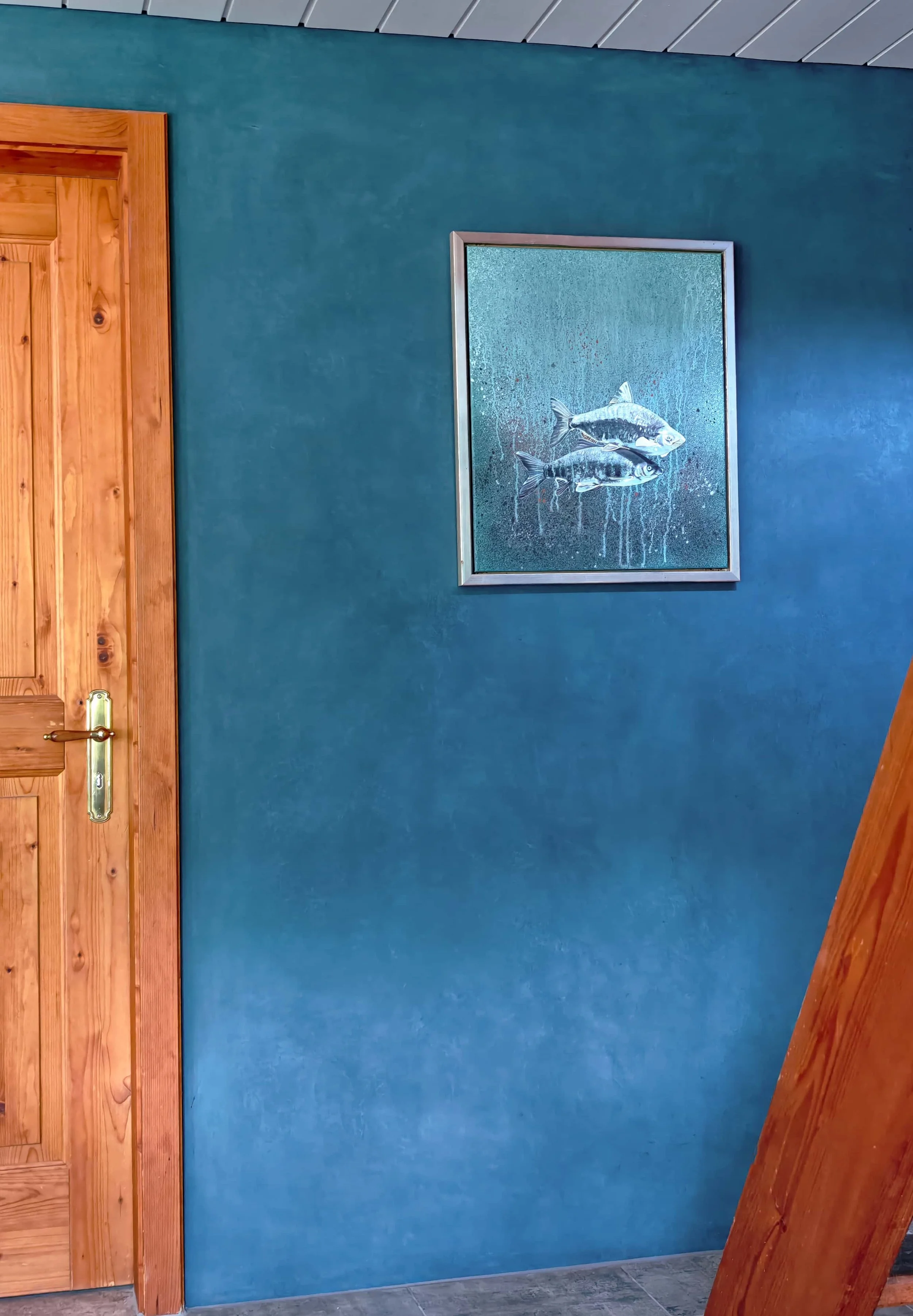 Blaue Wand mit einem Bilderrahmen mit einem Gemälde von zwei Fischen. Ein Holzrahmen und eine Tür sind ebenfalls sichtbar.