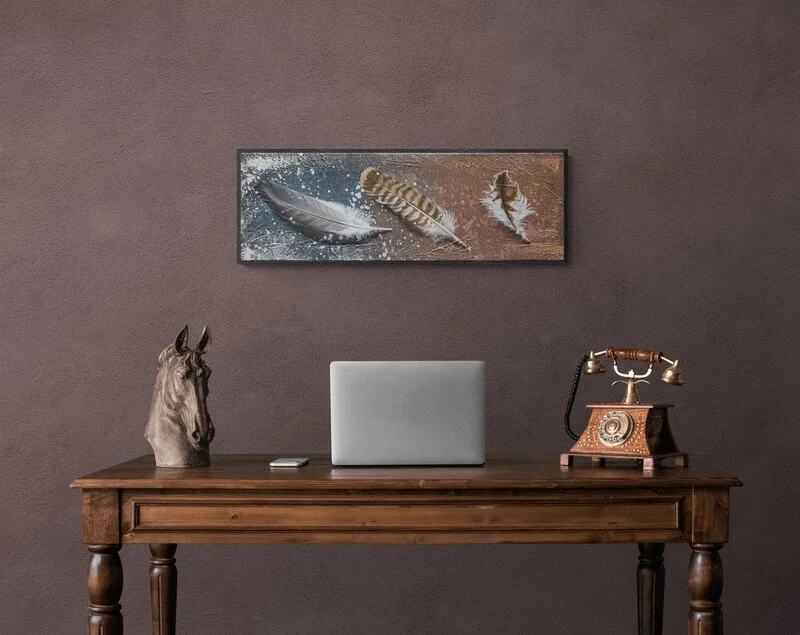 Ein moderner Büroarbeitsplatz mit einem Holzschreibtisch, einem Laptop, einem antiken Telefondrehgerät und einer Makrofotografie von drei Federn an der Wand.