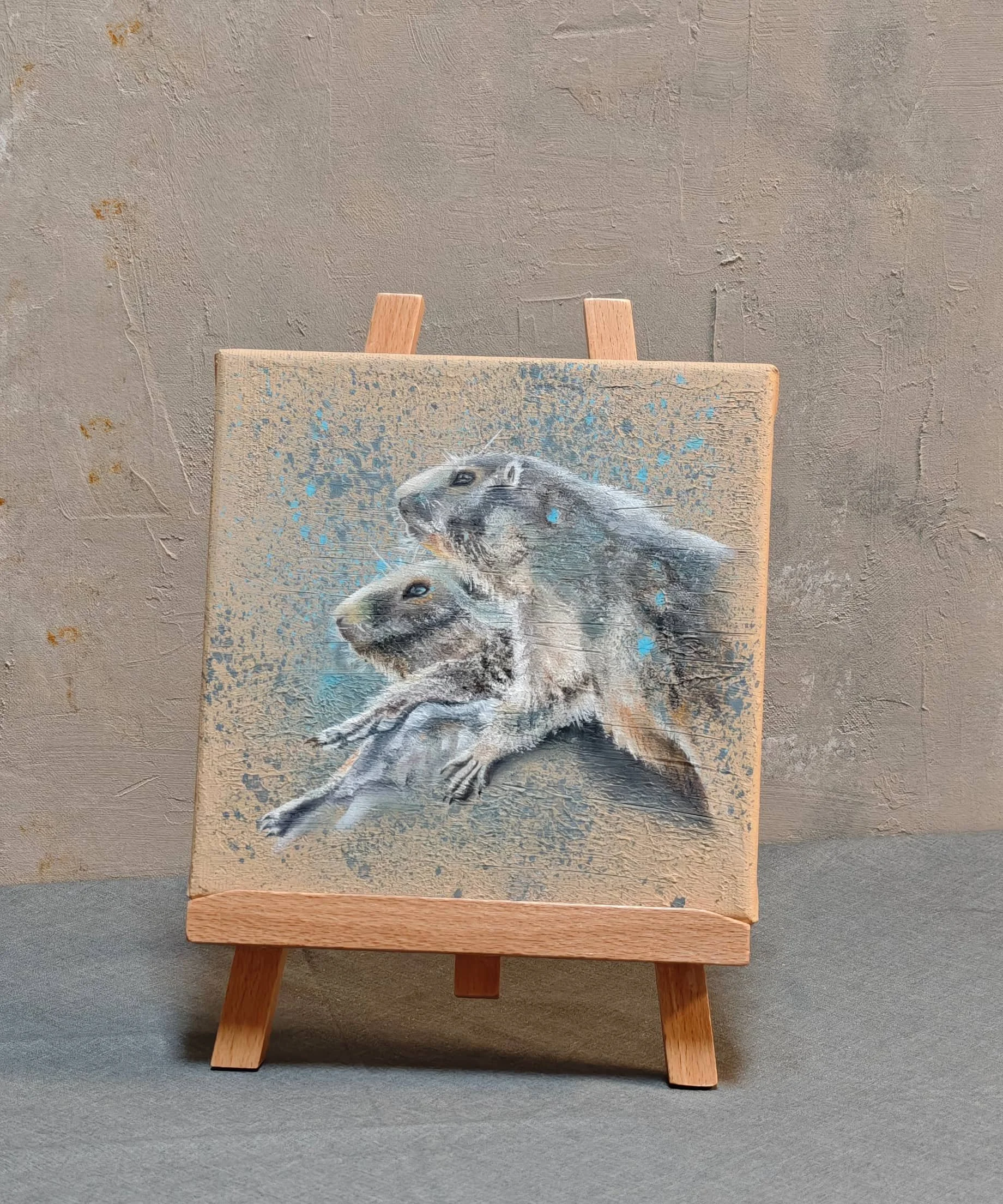 Kleines Gemälde auf einer Leinwand, das zwei streunende Hasen zeigt, mit blauem Akzent, auf einem kleinen Holzständer, vor einer rauen, hellgrauen Wand.