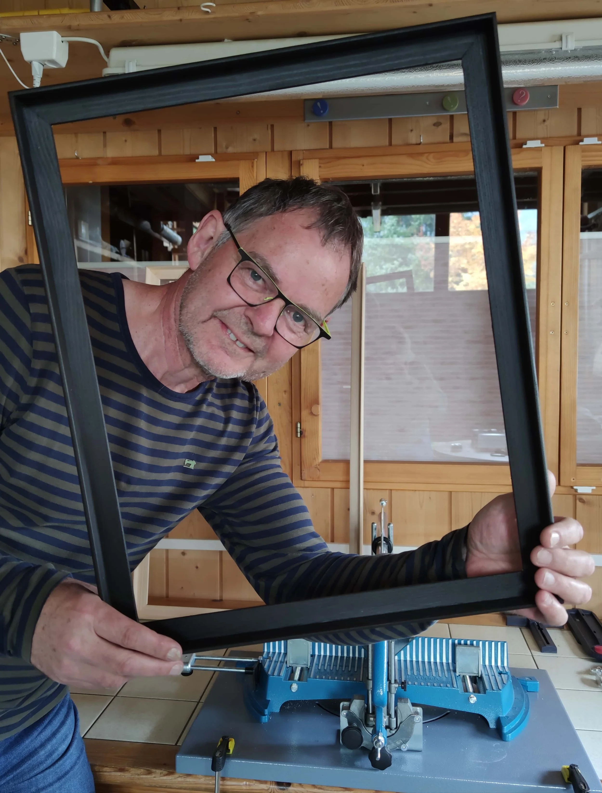 Ein Mann mit Brille und gestreiftem Pullover hält einen leeren Bilderrahmen und arbeitet an einem Werkstück in einer Werkstatt.