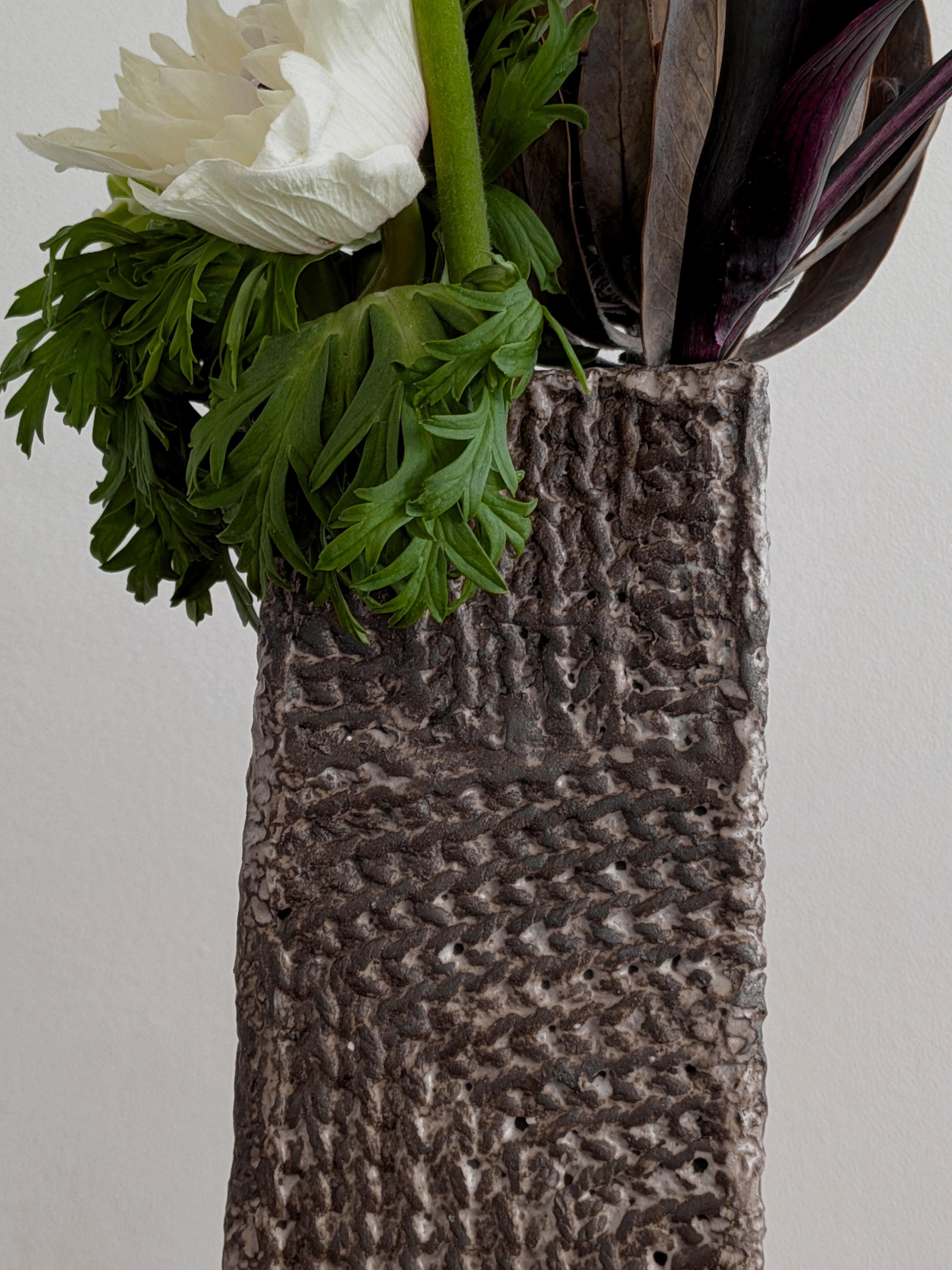 Vase en grès produit par Chloé Hercé contenant des plantes vertes, dont une grande feuille blanche, attaché à un fond blanc.