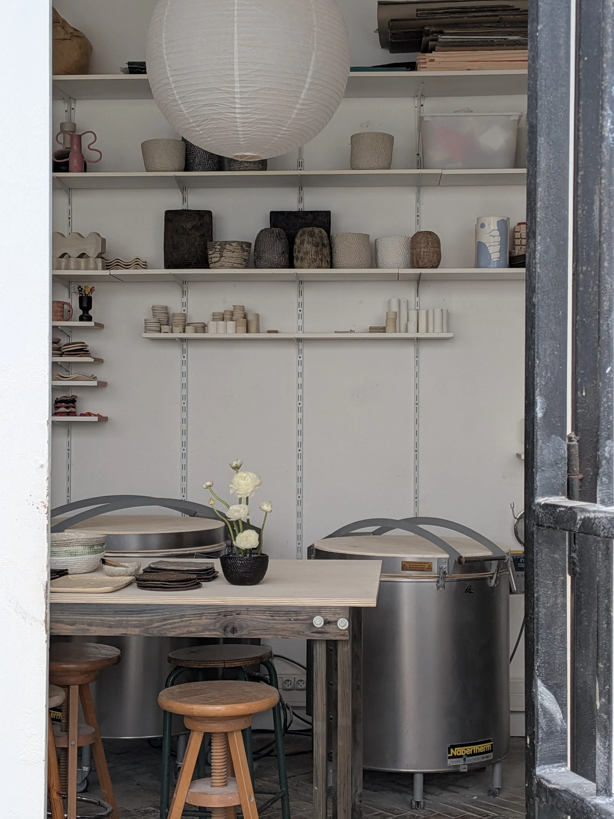 Vue de l'atelier de Chloé Hercé à Paris, avec des étagères contenant plusieurs pots et objets en céramique, un lampion en papier, une table avec des plateaux, des bouquets de fleurs blanches, des tabourets en bois.