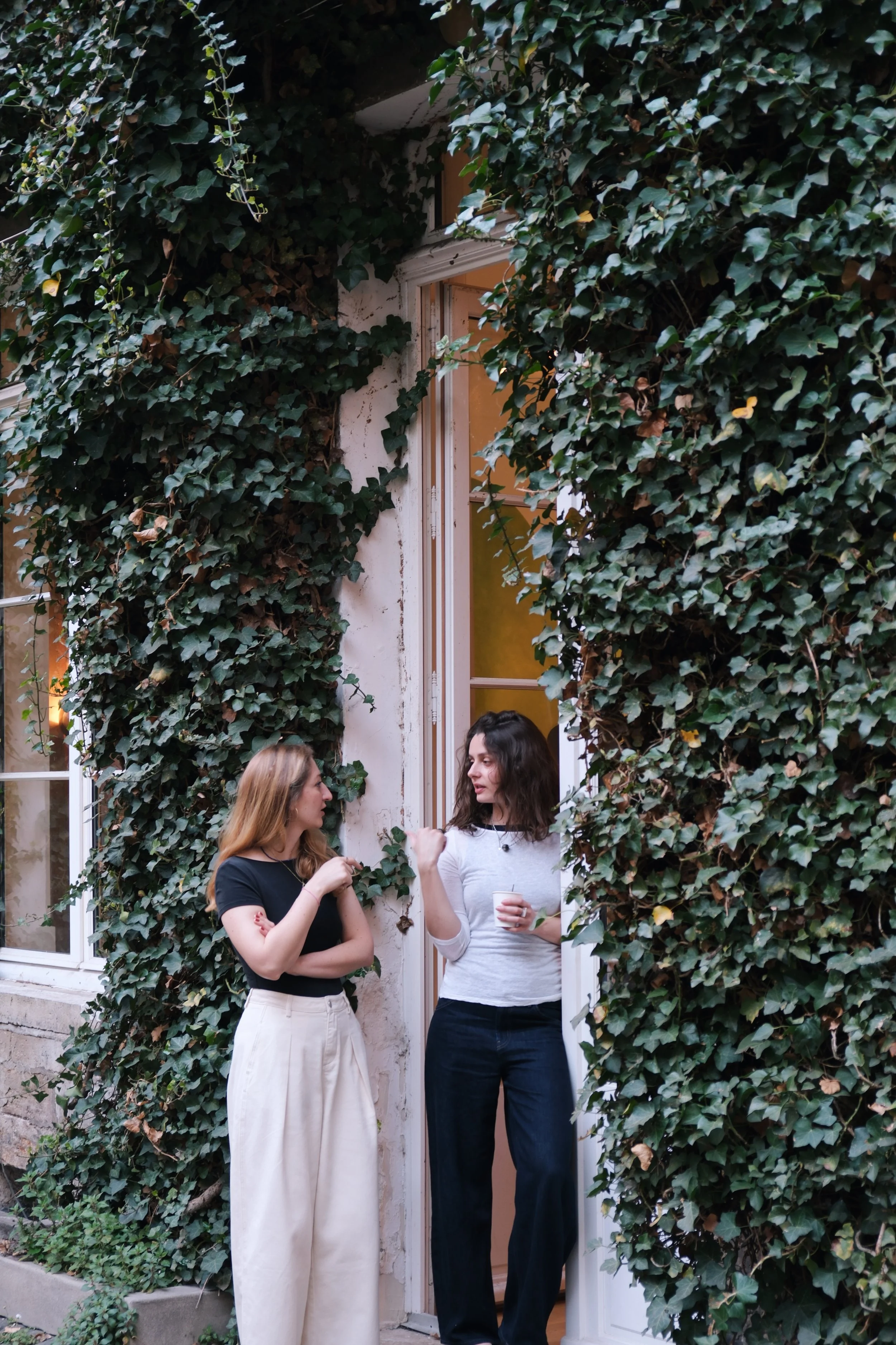 Chloé Hercé discute avec une artiste devant l'atelier parisien de la peintre d'Anna Peter Breton recouverte de feuilles de lierre, dans une ambiance chaleureuse.